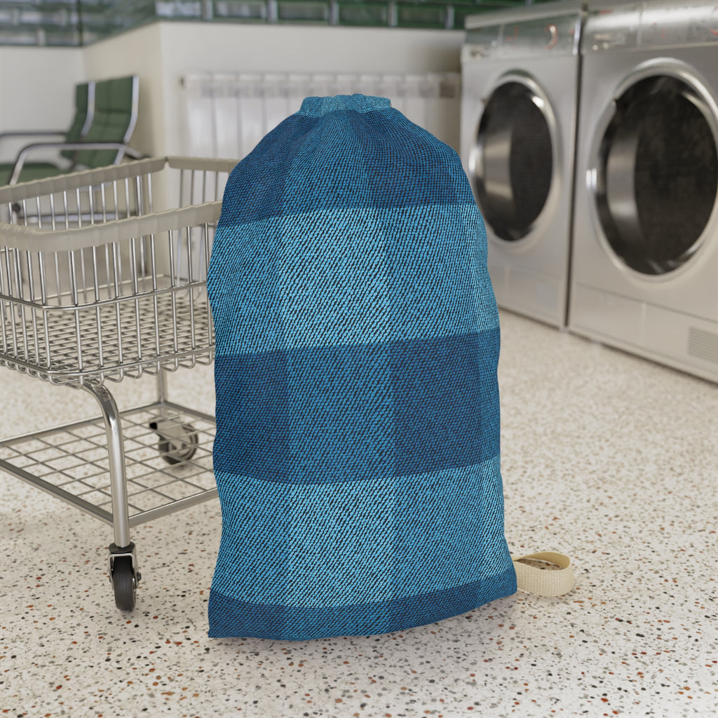 A large drawstring storage bag with a blue and teal buffalo plaid pattern, standing on a speckled laundry room floor next to a metal cart with washing machines in the background.