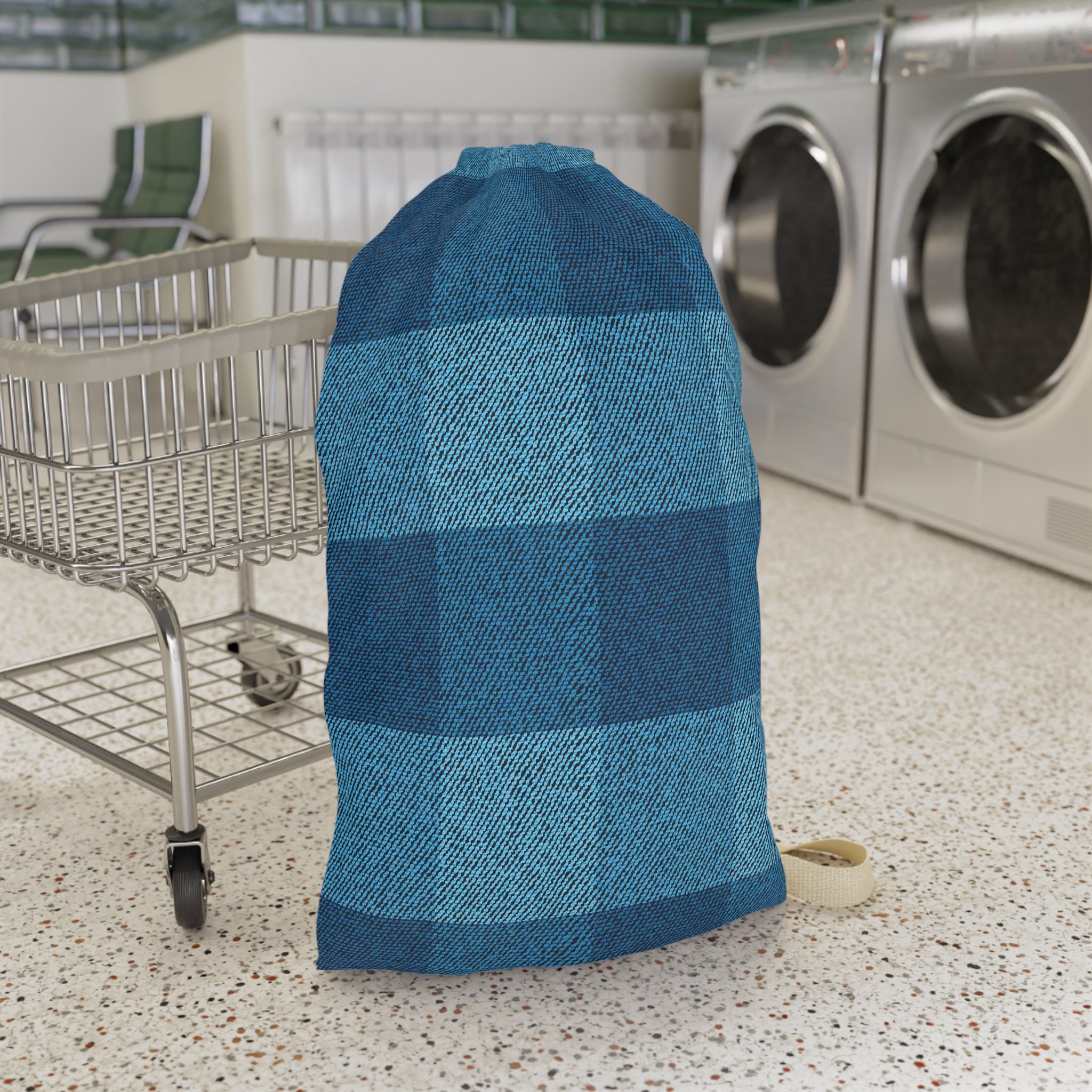 A large drawstring storage bag with a blue and teal buffalo plaid pattern, standing on a speckled laundry room floor next to a metal cart with washing machines in the background.