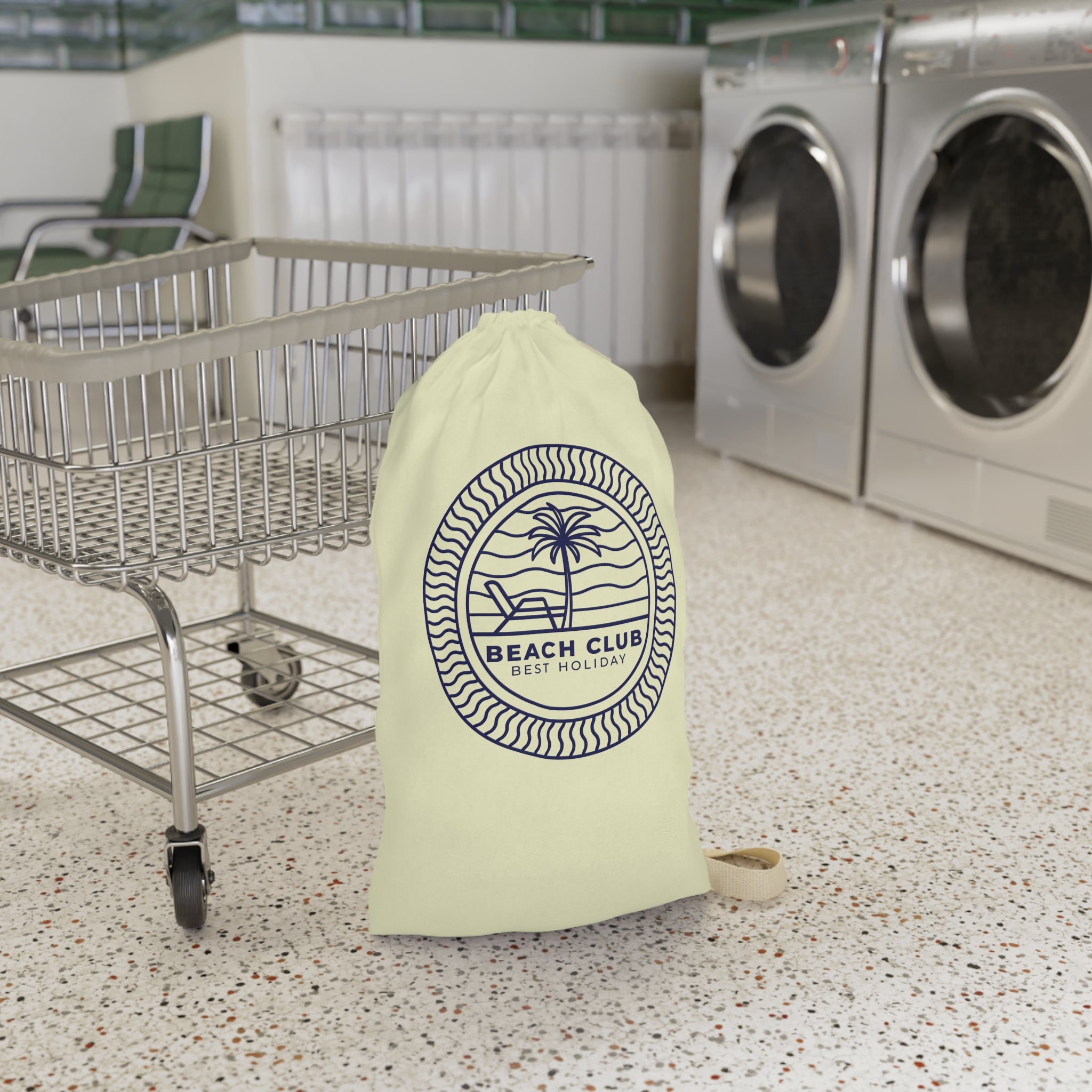 A pale yellow drawstring laundry bag with a navy blue 'Beach Club Best Money' palm tree logo, standing on a speckled floor next to a metal shopping cart in front of a laundry room with washing machines.