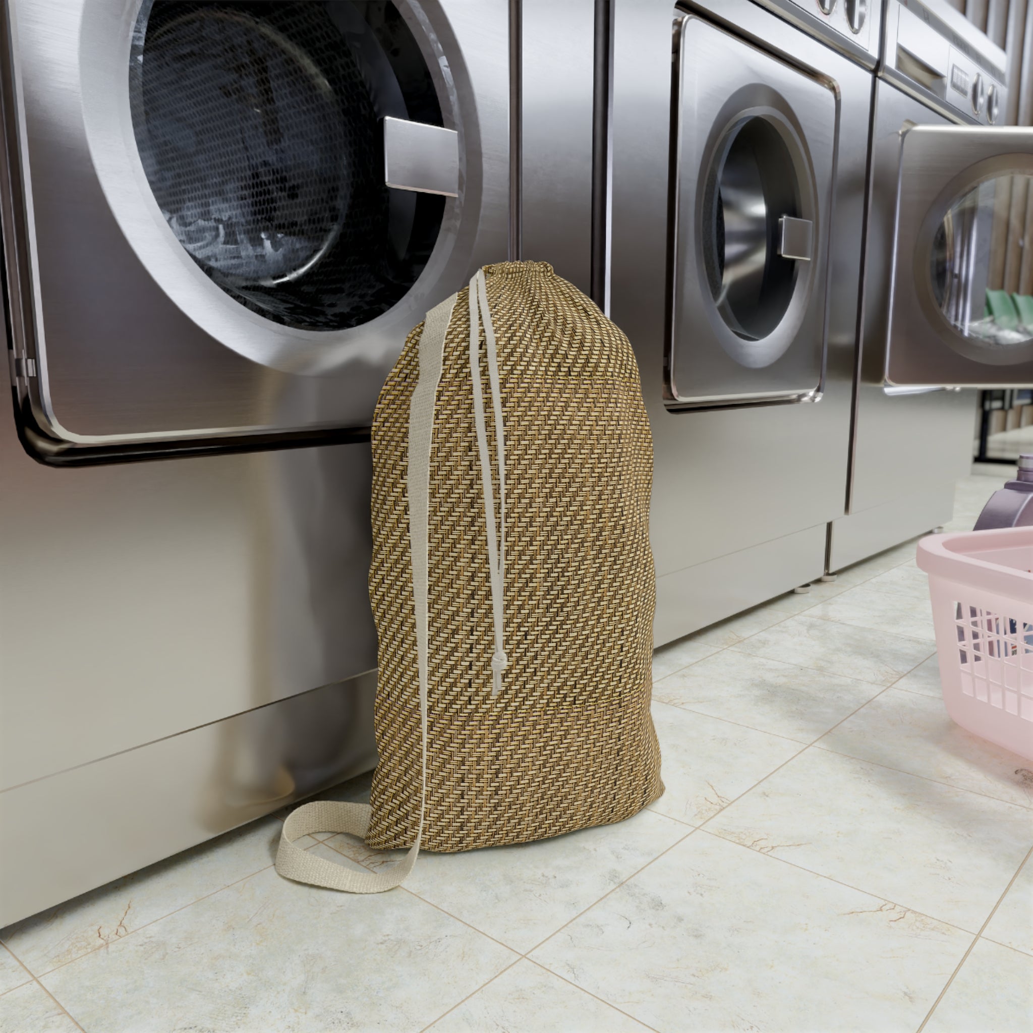 A woven beige laundry bag with white drawstrings stands on a tiled floor in front of stainless steel washing machines in a laundry room.