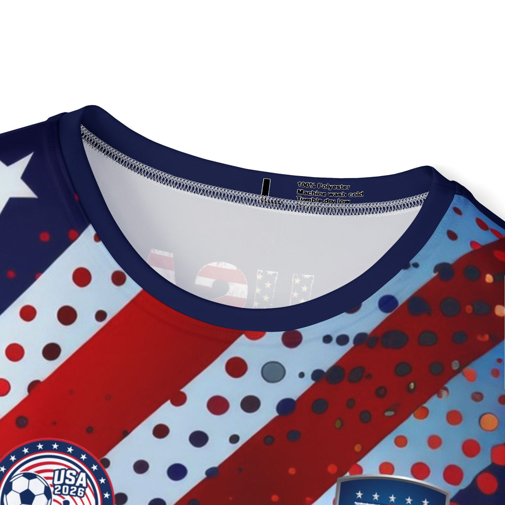 Close-up of a patriotic soccer jersey with red and light blue diagonal stripes, navy blue trim, and red, white, and blue polka dots. A circular 'USA 2026' patch is visible on the lower left.