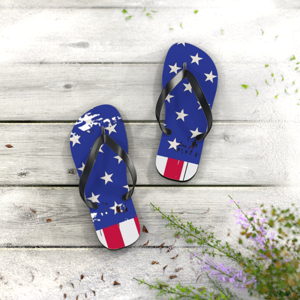 A pair of flip-flop sandals with a distressed American flag design, featuring white stars on a blue background and red and white stripes at the toe, displayed on a light gray wooden plank surface with decorative greenery.