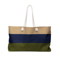 A horizontal-striped tote bag with white rope handles, featuring three color bands: beige on top, navy blue in the middle, and olive green on the bottom, with two white decorative pom-pom accents on the handles.