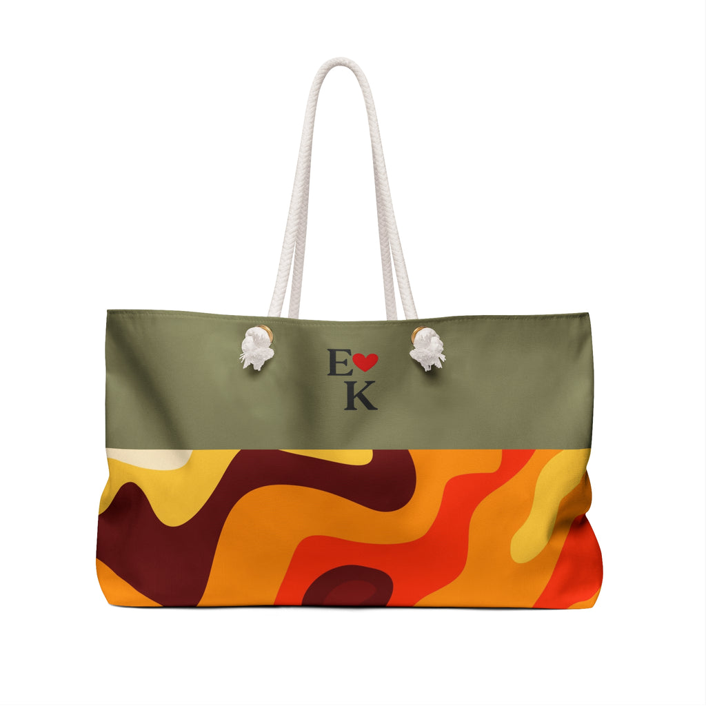 A tote bag with a two-tone design: the top half is olive green with white rope handles and a black 'E' and 'K' with a red heart printed in the center, while the bottom half features an abstract wavy pattern in orange, yellow, red, and dark brown.