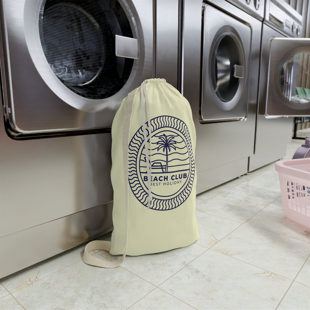 A cream-colored drawstring laundry bag with a navy blue 'Beach Club' palm tree logo, standing on a tiled floor in front of stainless steel washing machines.