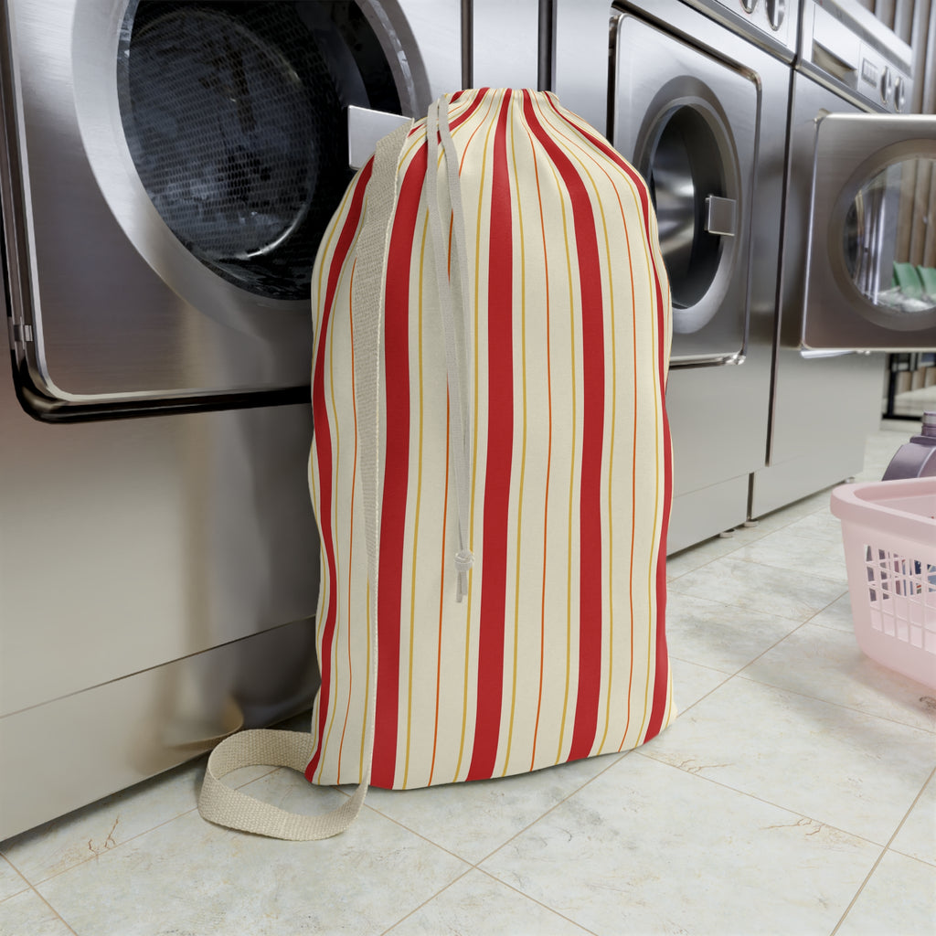 A striped laundry bag with red, cream, and thin yellow vertical stripes, featuring a cream zipper and strap, standing on a tiled floor in front of stainless steel washing machines.