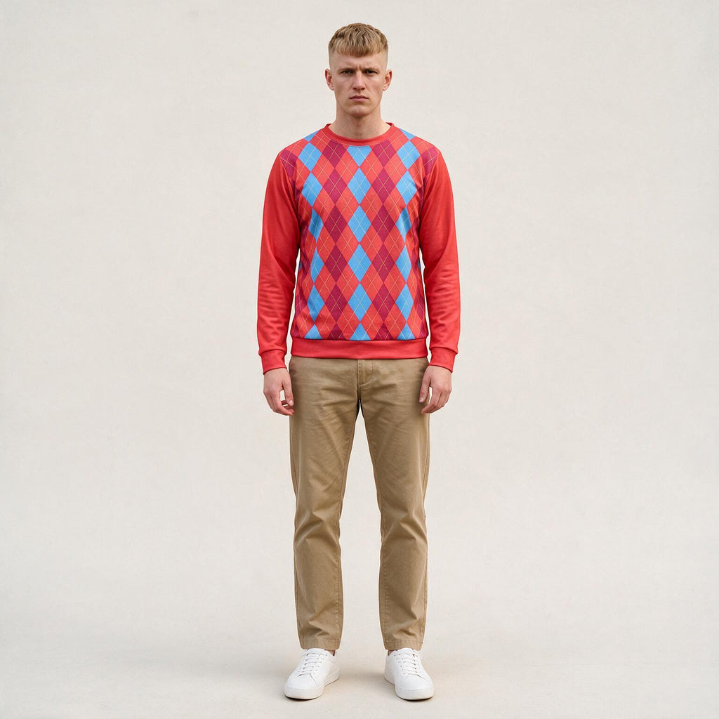 A man stands against a plain light beige studio background wearing a long-sleeve crewneck sweatshirt with a red, pink, and blue argyle pattern, khaki pants, and white sneakers.
