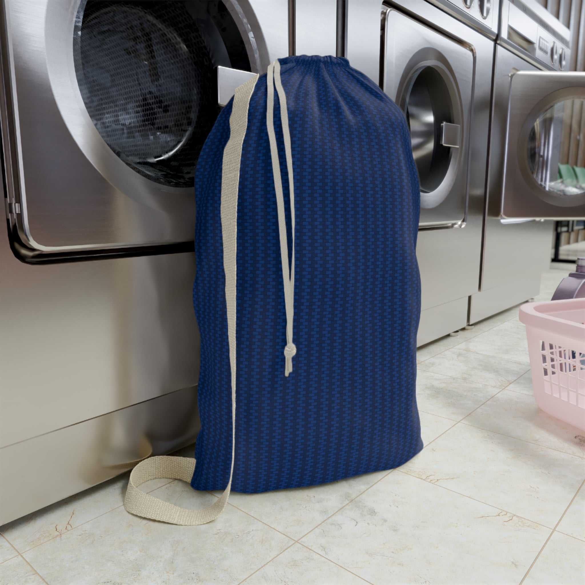 A blue drawstring laundry bag with a subtle geometric pattern, standing on a tiled floor in front of stainless steel washing machines.