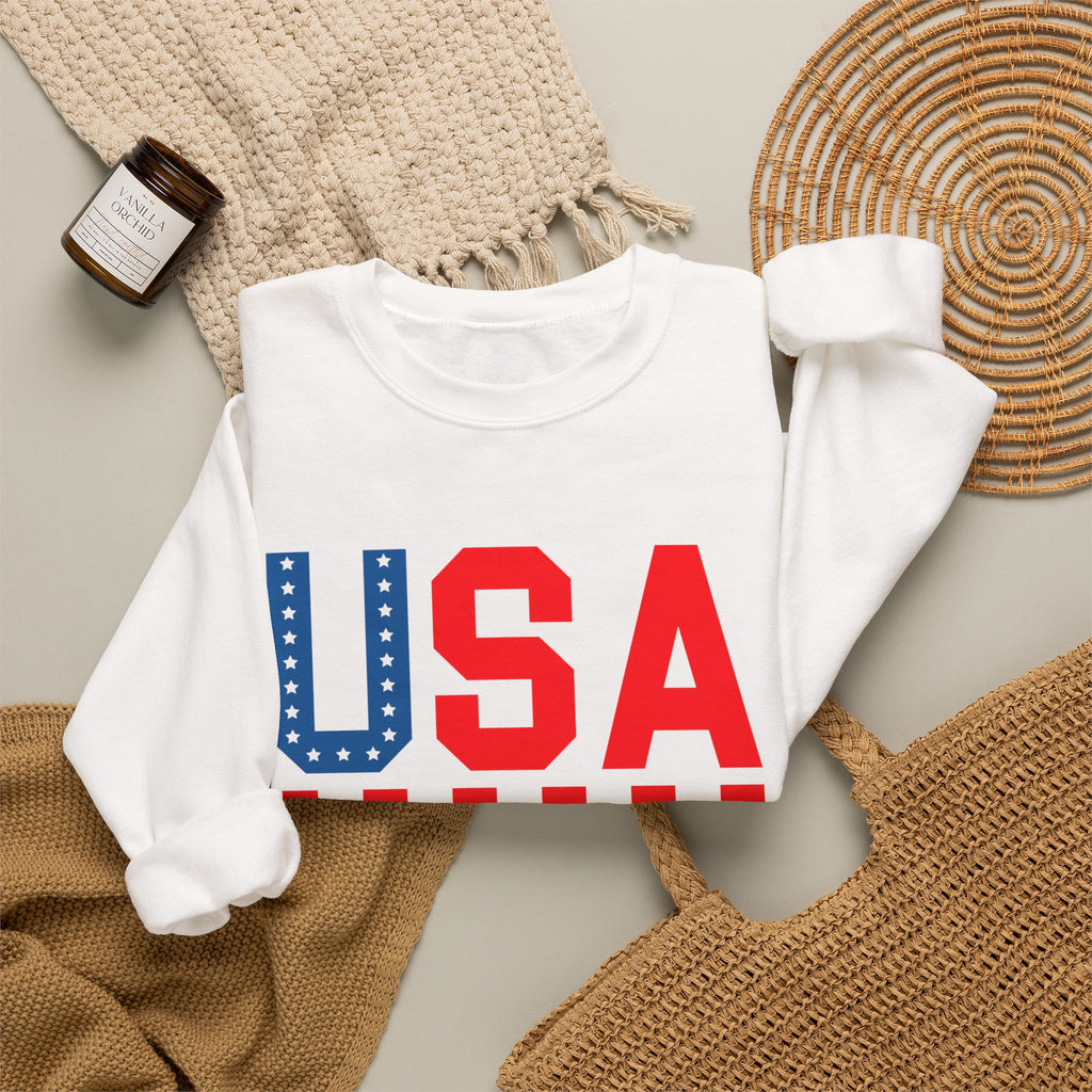 A folded white long-sleeve sweatshirt with a large 'USA' graphic across the chest, styled in a flat lay with a woven blanket, a small candle jar, a wicker tray, and a tan woven bag on a light neutral background.