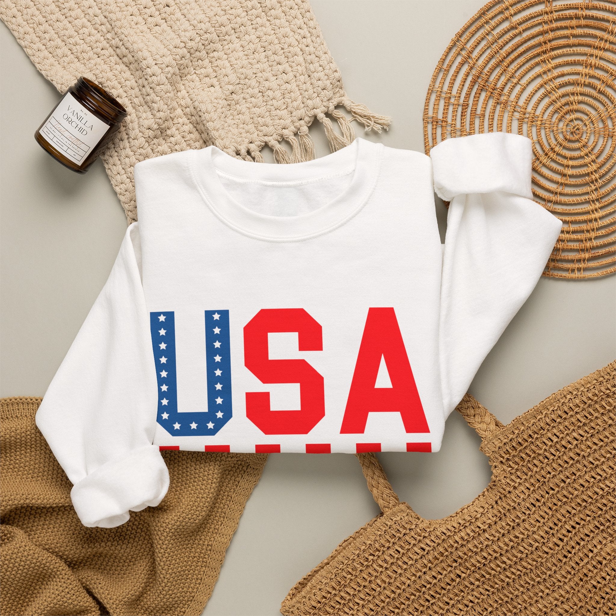 A folded white long-sleeve sweatshirt with a large 'USA' graphic across the chest, styled in a flat lay with a woven blanket, a small candle jar, a wicker tray, and a tan woven bag on a light neutral background.