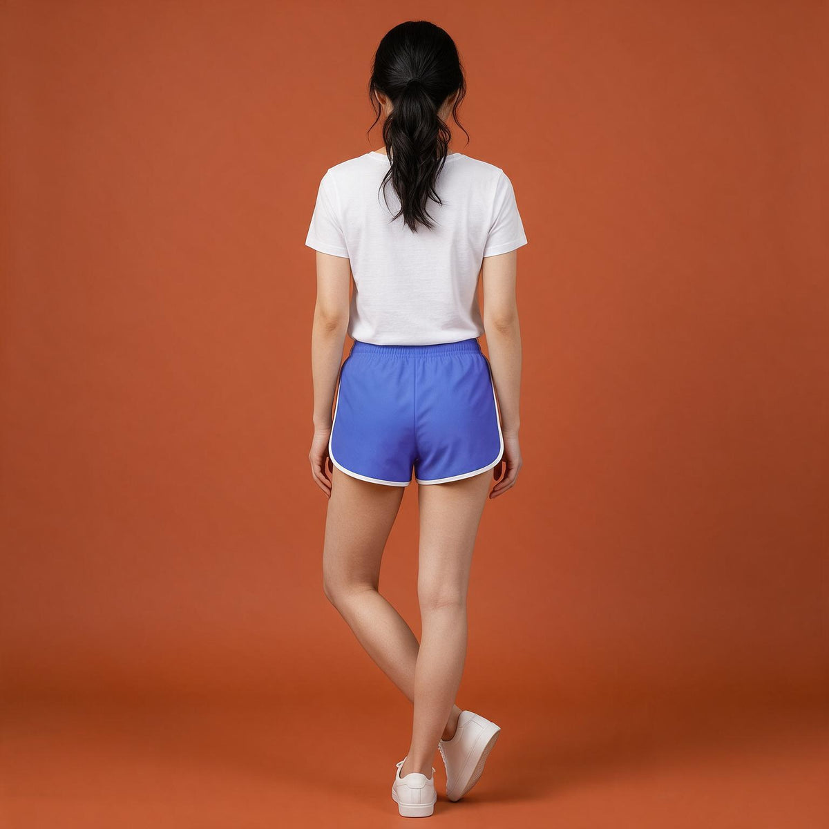 A woman is shown from behind wearing blue athletic shorts with white trim, a white t-shirt, and white sneakers, standing against a solid orange studio background.