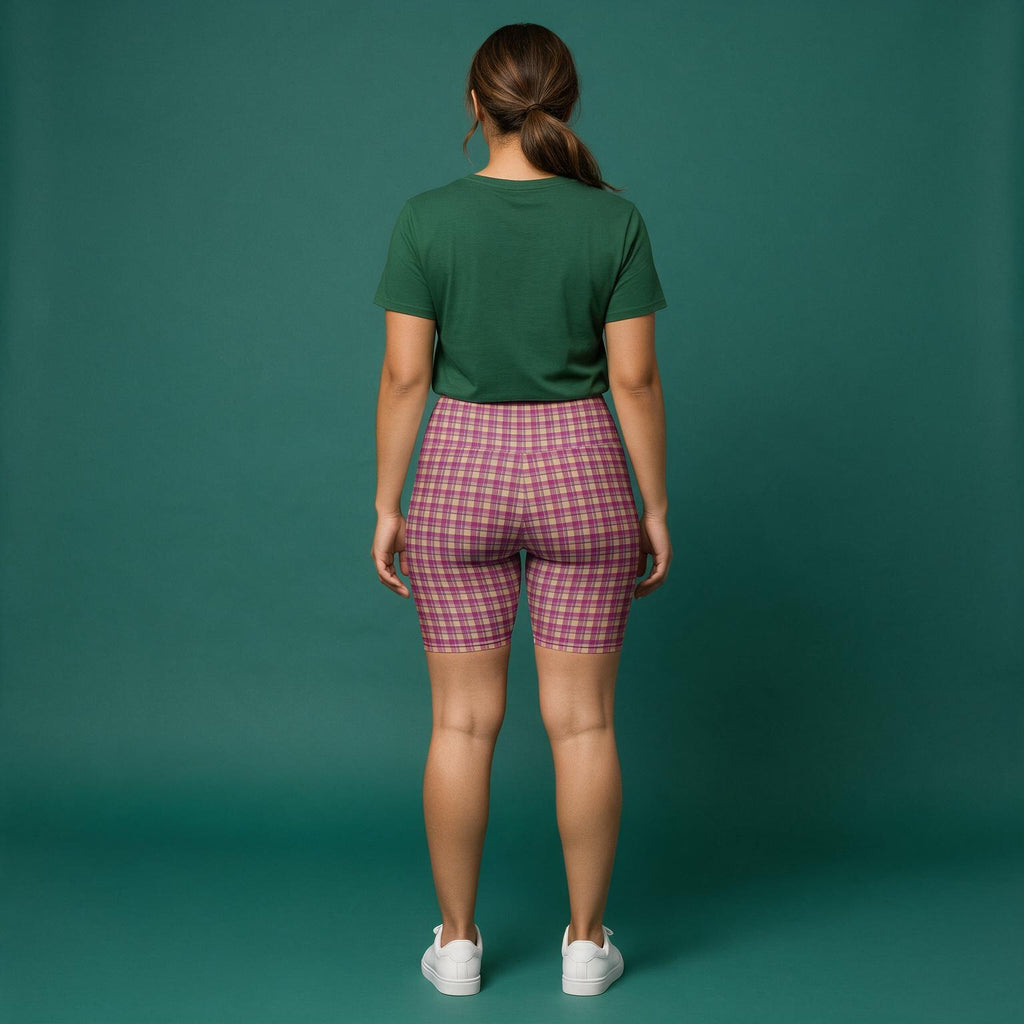 A woman is shown from behind wearing pink and purple plaid biker shorts, a dark green t-shirt, and white sneakers against a solid teal-green studio backdrop.