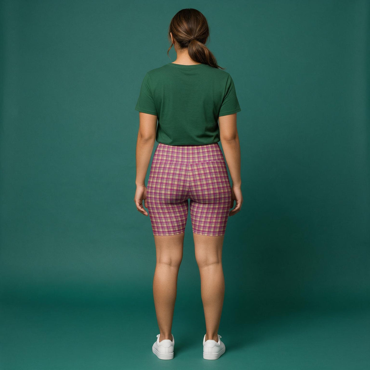 A woman is shown from behind wearing pink and purple plaid biker shorts, a dark green t-shirt, and white sneakers against a solid teal-green studio backdrop.