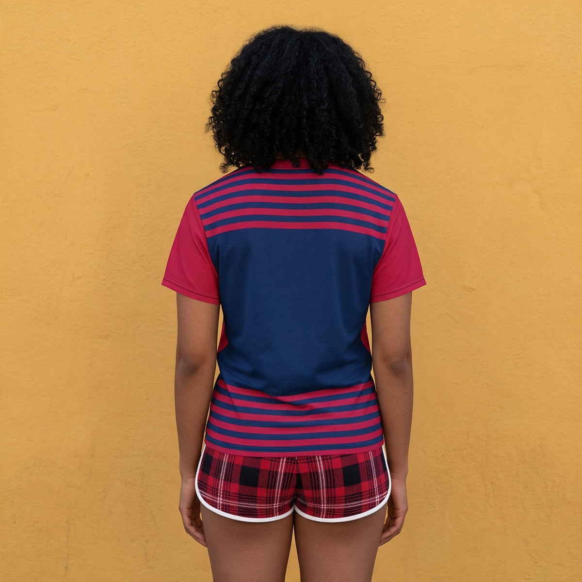 A person viewed from behind wearing a short-sleeve athletic top with a navy blue back panel and red-and-navy horizontal stripes on the shoulders and lower back, paired with red plaid shorts with white trim, standing against a solid mustard-yellow wall.