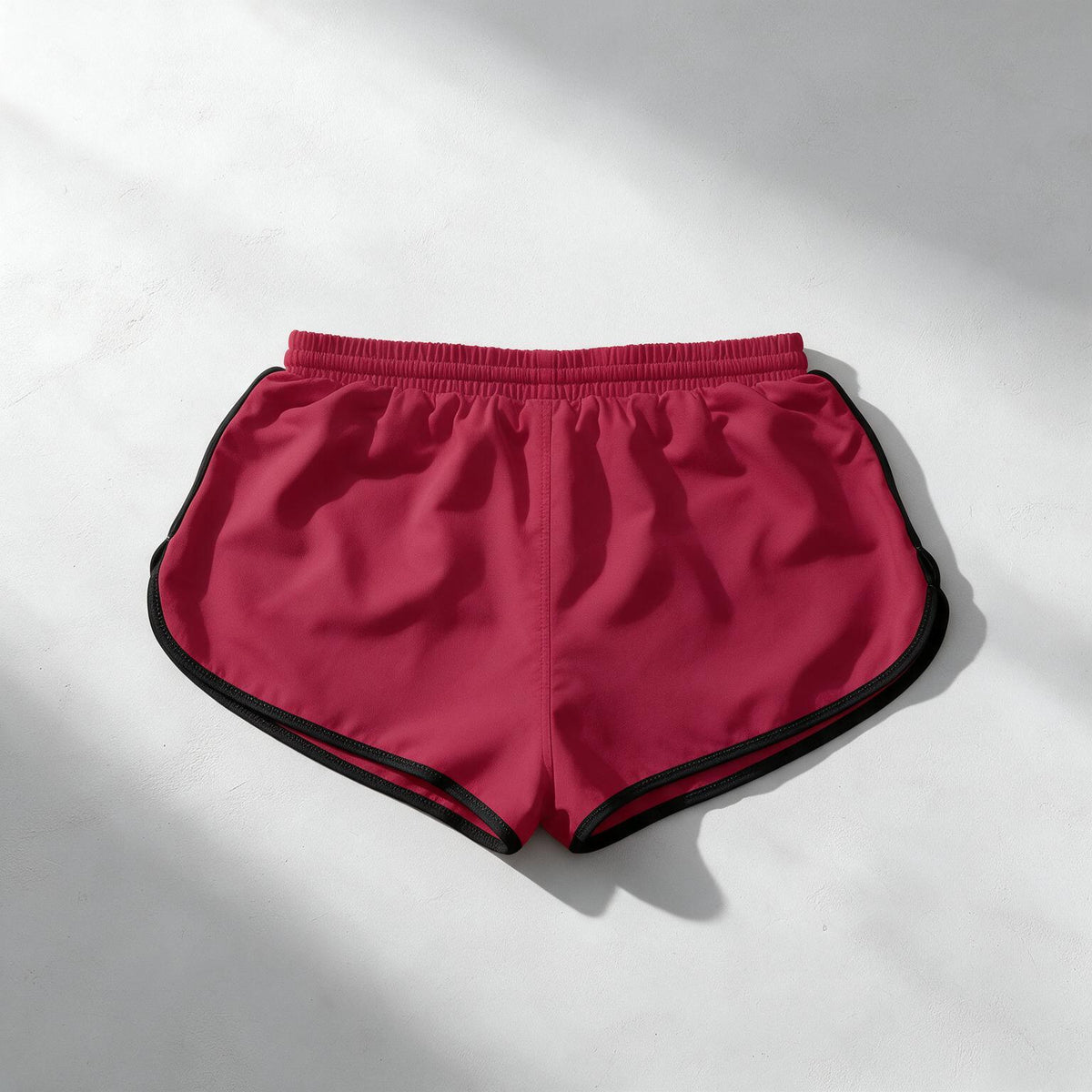 A pair of athletic shorts in a deep magenta pink with black piping along the edges, laid flat on a light gray surface with soft diagonal shadows.
