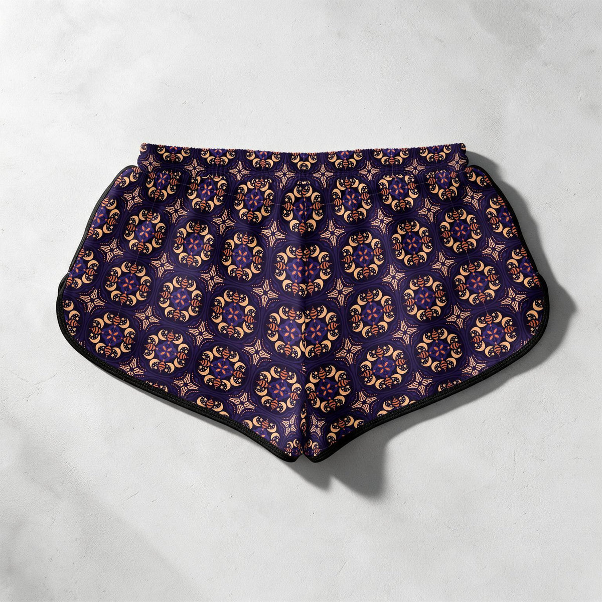 A pair of women's athletic shorts featuring an all-over geometric medallion pattern in purple, gold, and beige, with black trim and an elastic waistband, laid flat on a light gray textured surface.