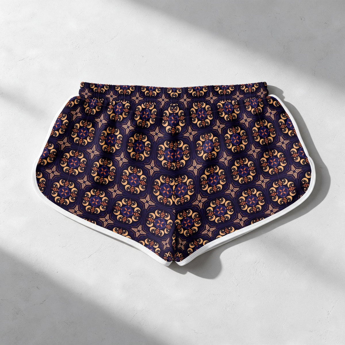 Patterned athletic shorts with a dark navy base, featuring an intricate gold and purple mandala pattern, and a contrasting white side trim, laid flat on a light gray textured surface with soft shadows.