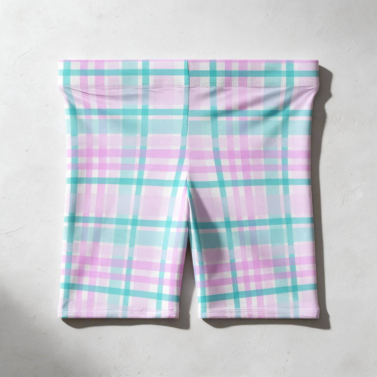 Pink and teal plaid biker shorts laid flat on a light gray textured background.