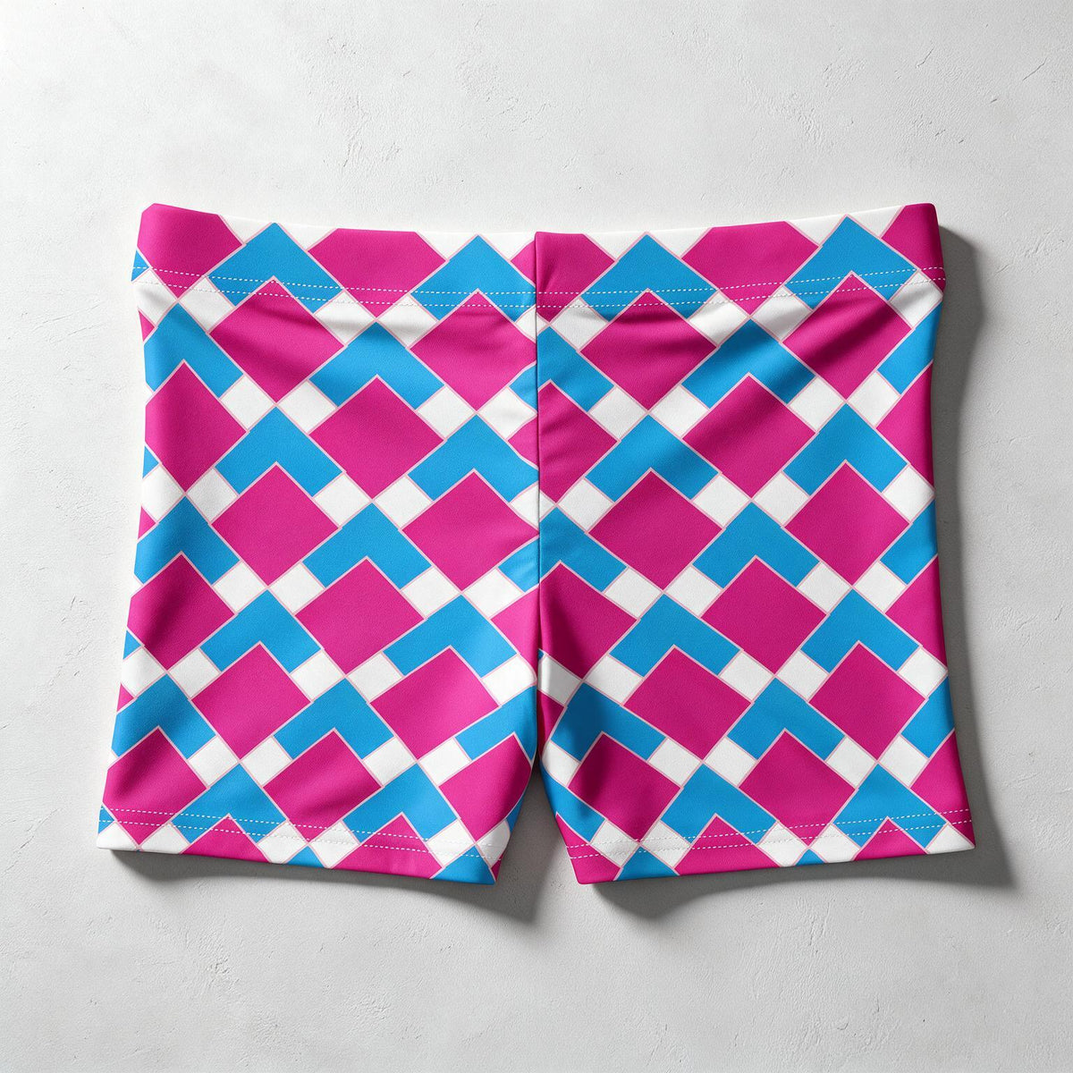 A pair of athletic shorts featuring a bold geometric pattern of magenta pink, bright blue, and white interlocking diamond shapes, laid flat on a light gray textured surface.