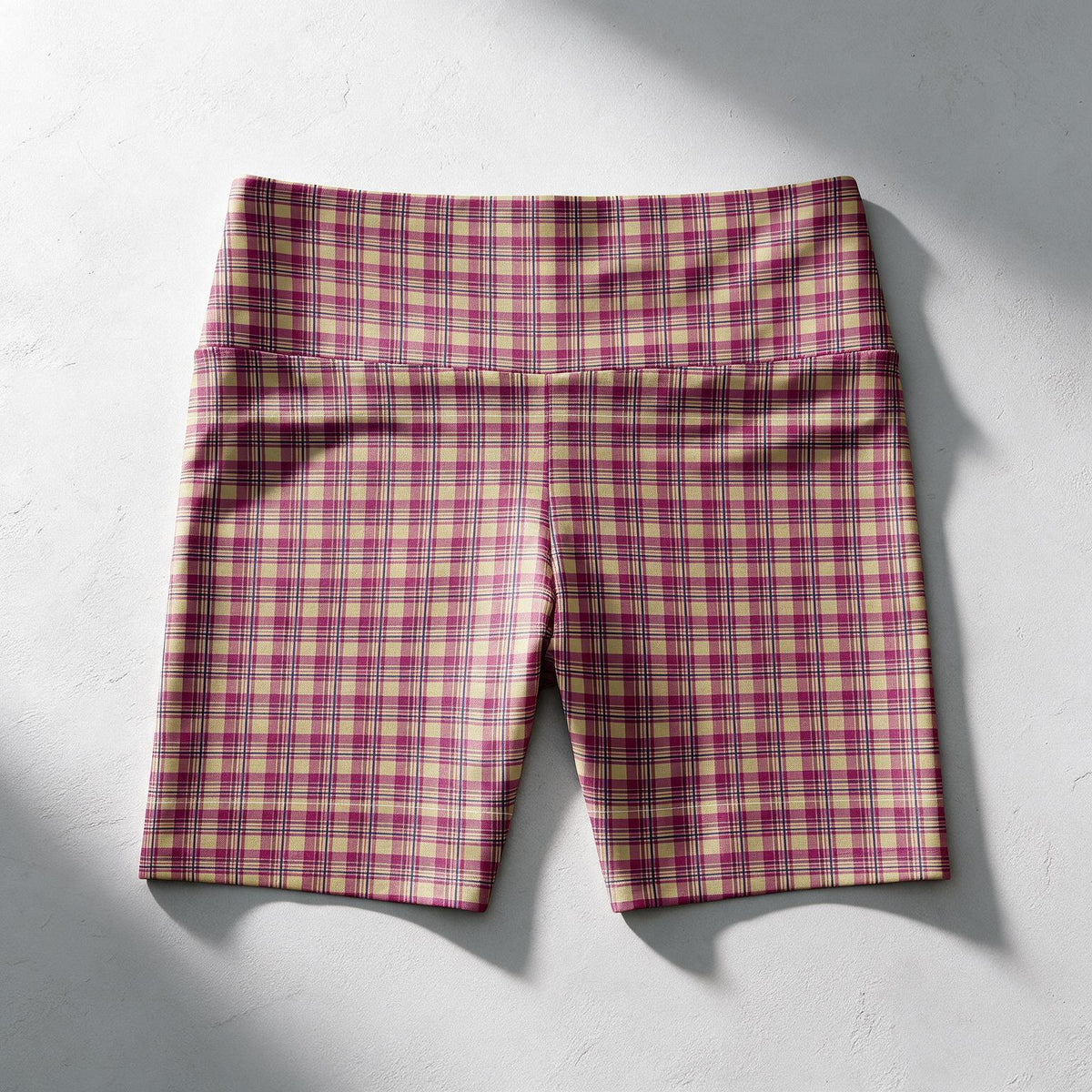 Plaid high-waisted bike shorts in a pink, purple, and beige pattern, laid flat on a textured light gray background with soft shadows.