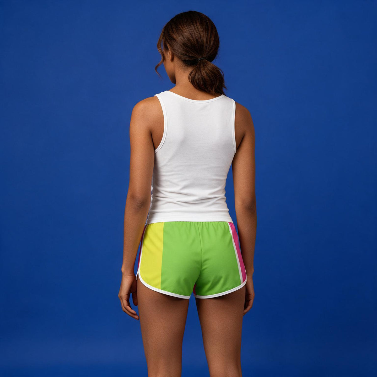 A woman shown from behind wearing bright color-blocked athletic shorts with green, yellow, and pink panels, paired with a white tank top, against a solid blue studio background.