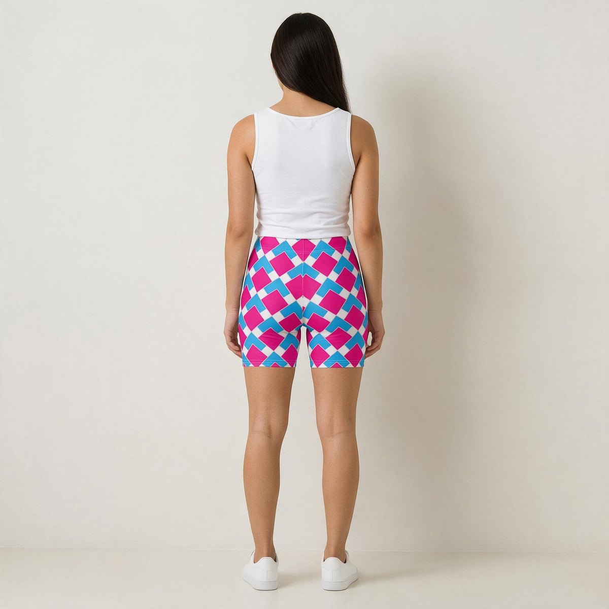 A woman stands with her back to the camera wearing bright geometric print athletic shorts with a pink, blue, and white diamond pattern, paired with a white tank top and white sneakers.