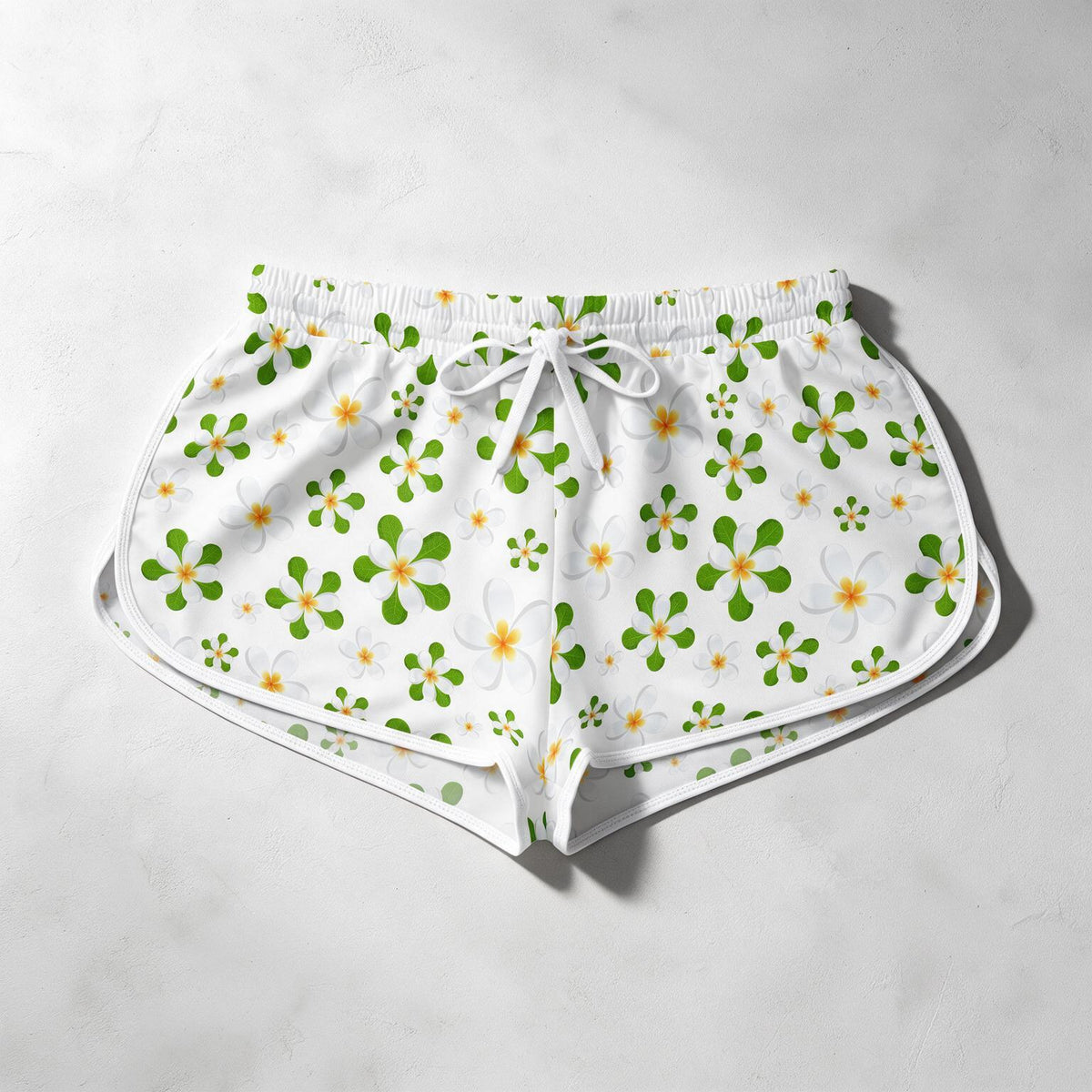 White shorts with a green and yellow tropical floral pattern, featuring a white drawstring waist, laid flat on a light gray textured background.