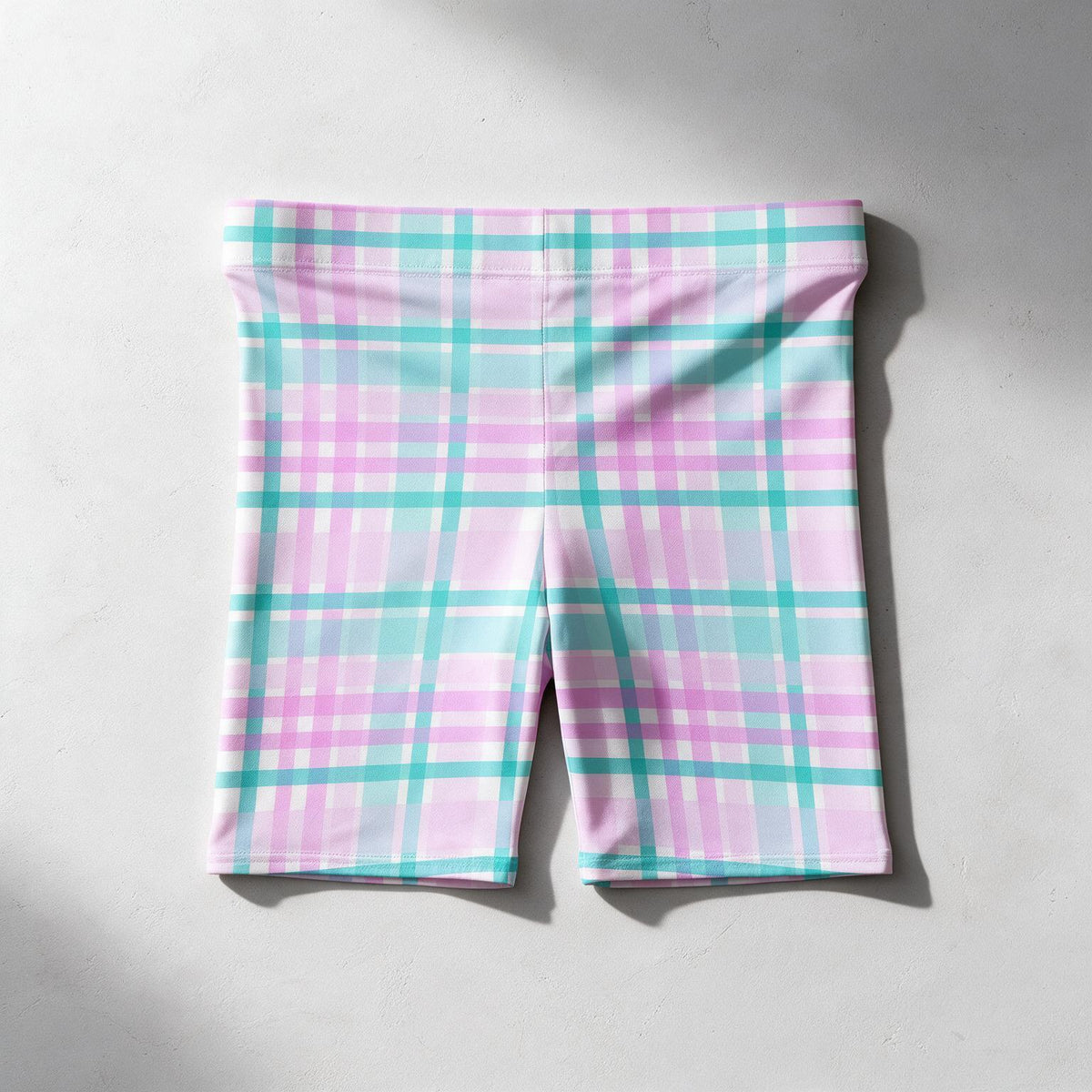 Pink and turquoise plaid athletic shorts laid flat on a light gray concrete surface.