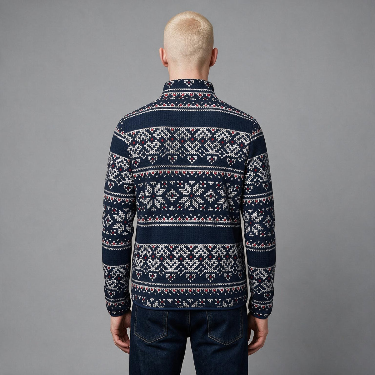 A man viewed from behind wearing a navy blue knit sweater with a white and red Nordic/Fair Isle pattern, paired with dark blue jeans, against a solid gray studio background.