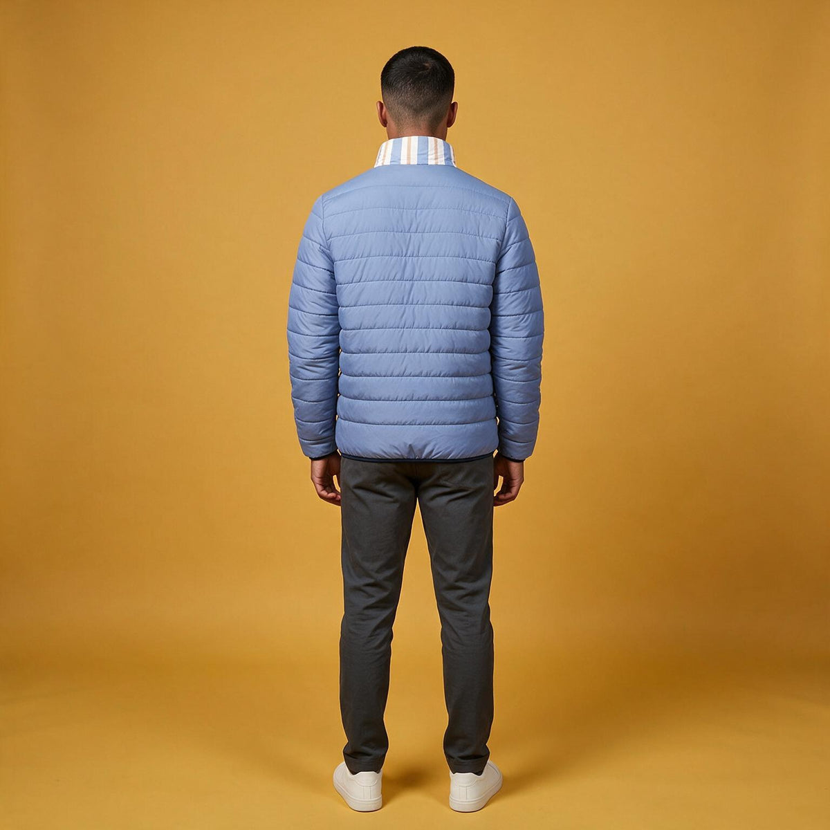 A man viewed from the back wearing a light blue puffer jacket, dark gray trousers, and white sneakers, standing against a solid mustard yellow studio background.
