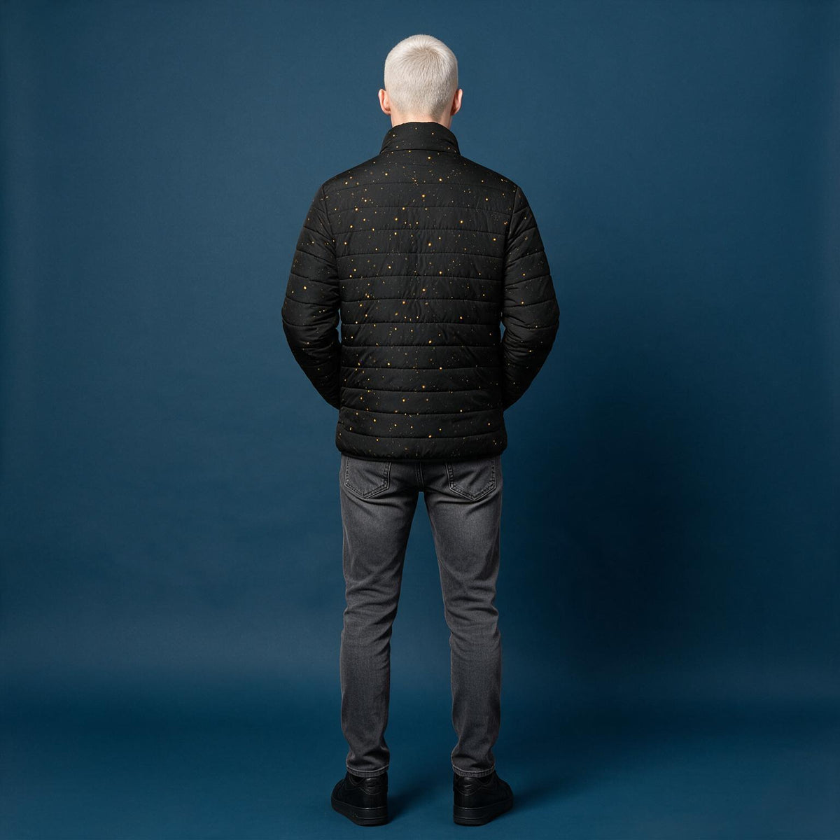 A man viewed from behind wearing a black quilted puffer jacket with small gold dot details, paired with gray jeans and black shoes, standing against a solid dark blue studio backdrop.