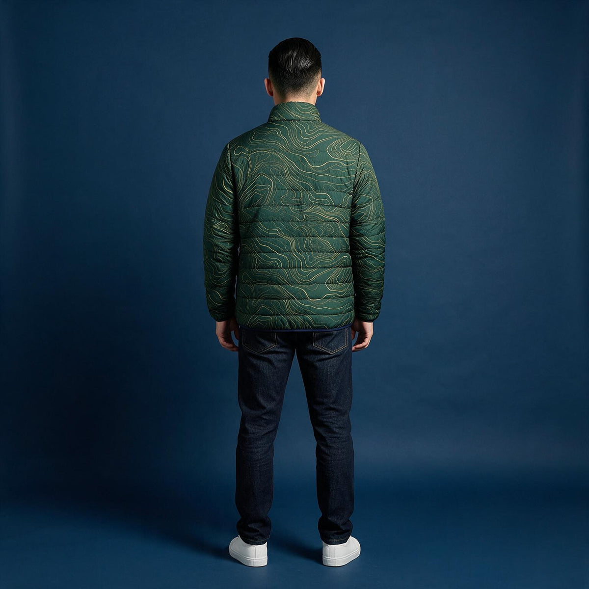 A man viewed from behind wearing a dark green quilted puffer jacket with a wavy topographic pattern, dark jeans, and white sneakers, standing against a dark blue studio backdrop.