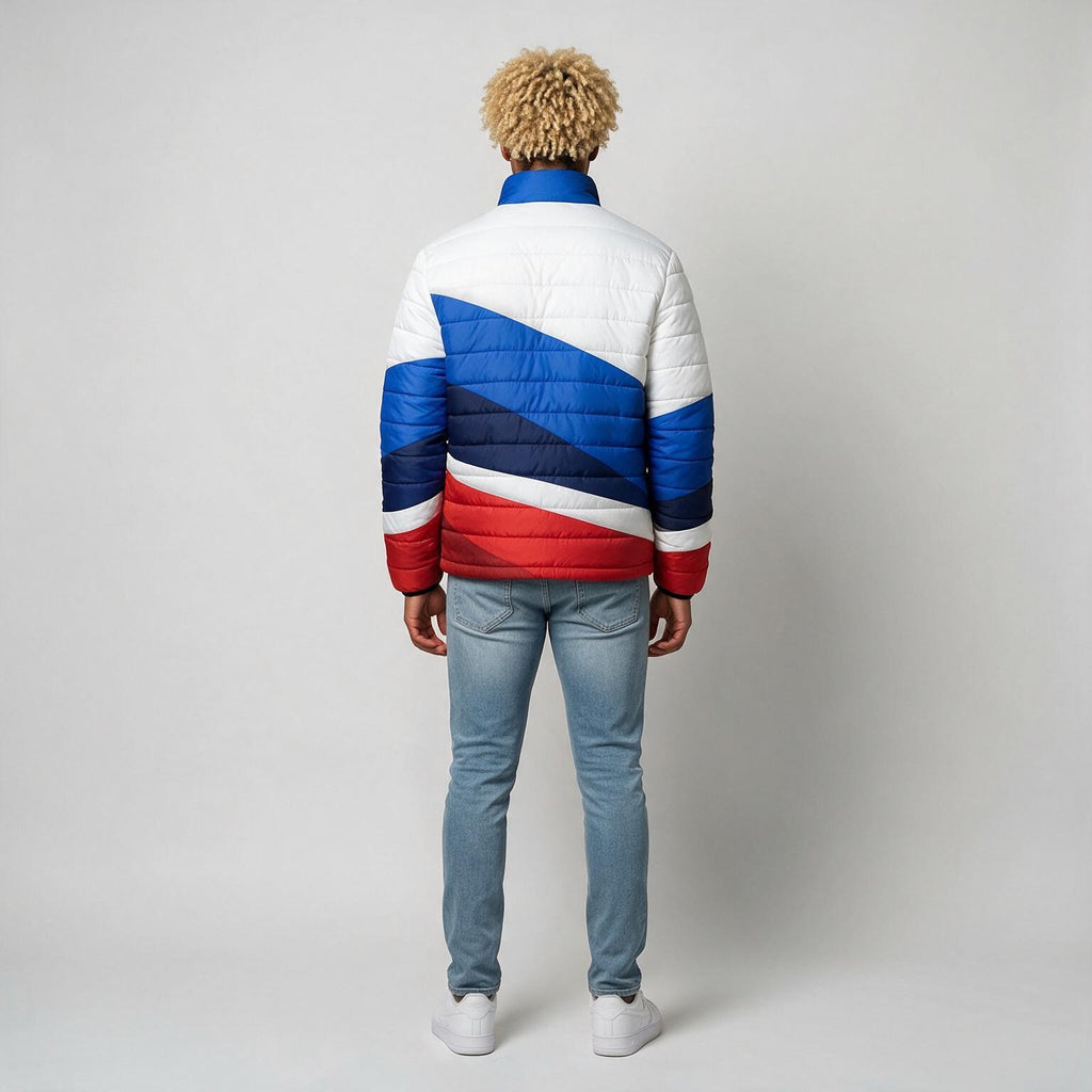 A man viewed from behind wearing a puffer jacket with a bold diagonal color-blocked pattern in white, blue, and red, paired with light blue jeans and white sneakers, against a plain light gray studio background.