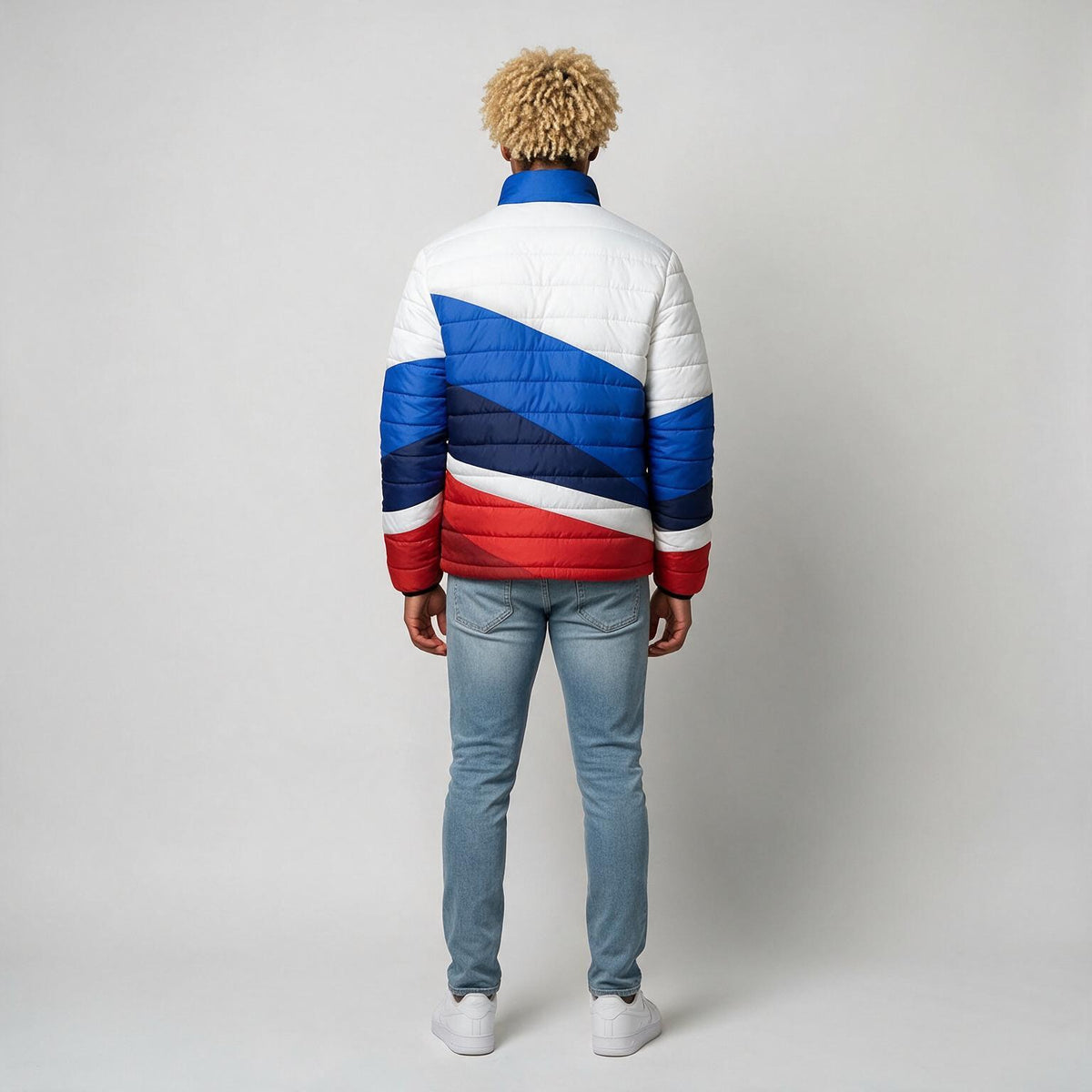 A man viewed from behind wearing a puffer jacket with a bold diagonal color-blocked pattern in white, blue, and red, paired with light blue jeans and white sneakers, against a plain light gray studio background.