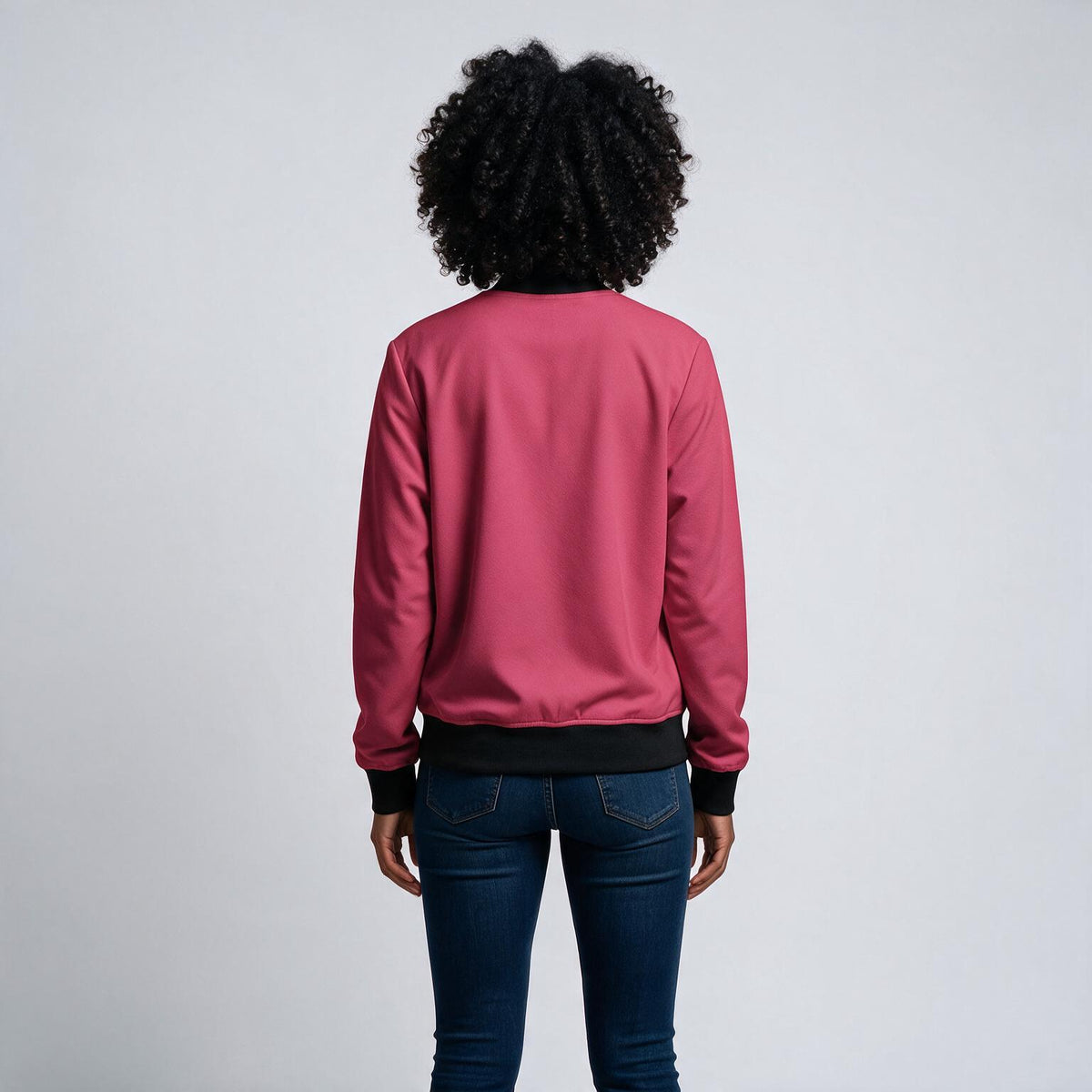 A person viewed from the back wearing a pink bomber-style sweatshirt with black ribbed trim and dark blue jeans, standing against a plain light gray studio background.