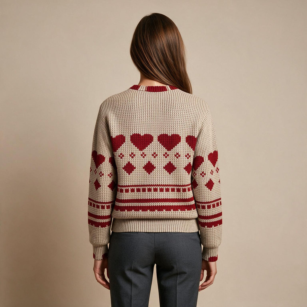 A woman viewed from behind wearing a cream-colored knit sweater with a red Nordic heart and diamond pattern, paired with dark gray trousers, against a solid beige background.