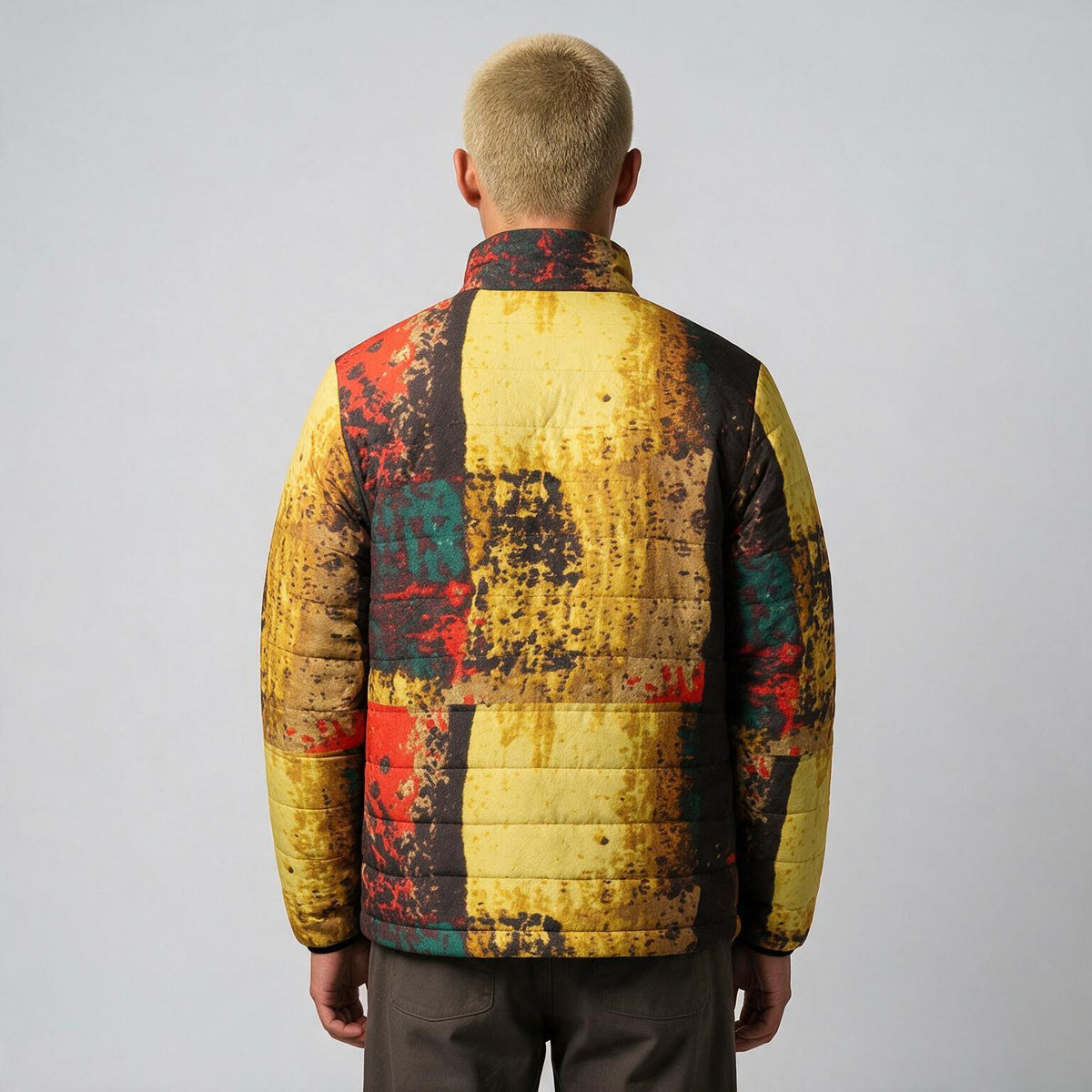 A man viewed from behind wearing a quilted puffer jacket with a distressed abstract patchwork pattern in yellow, red, black, and teal, set against a plain light gray studio background.