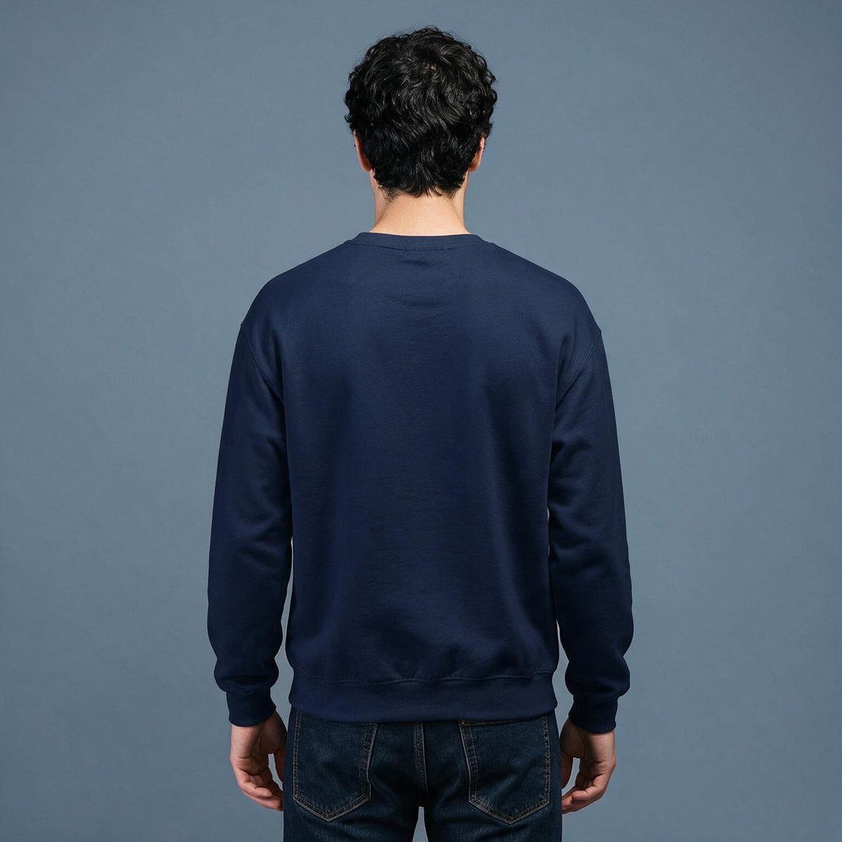 A man viewed from behind wearing a dark navy blue crewneck sweatshirt and dark jeans, standing against a solid slate blue-gray background.