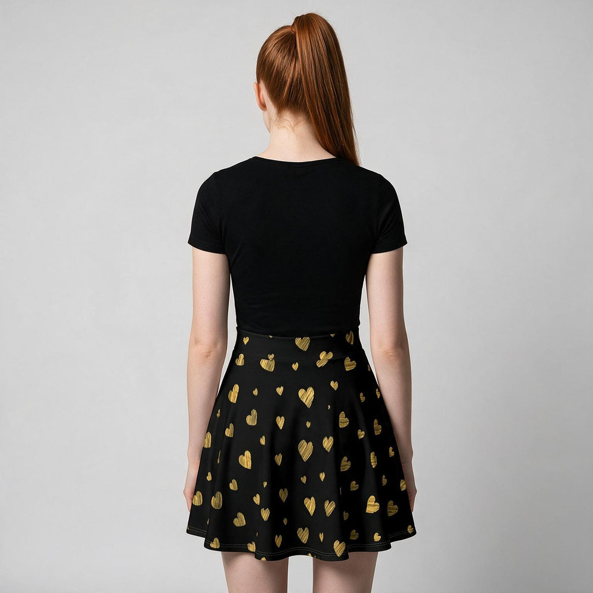 A woman viewed from behind wearing a black short-sleeve top and a black flared skirt with a repeating metallic gold heart pattern, against a light gray studio background.