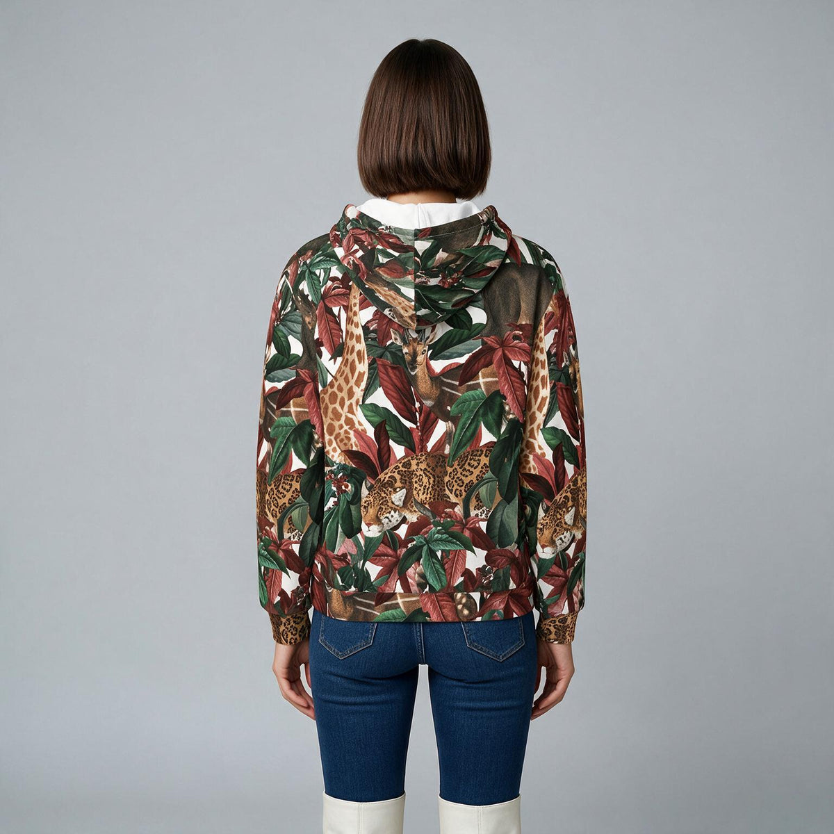 A model viewed from the back wearing a hooded sweatshirt with an all-over safari print of giraffes and leopards among tropical leaves, paired with blue jeans and white boots, against a solid gray studio background.
