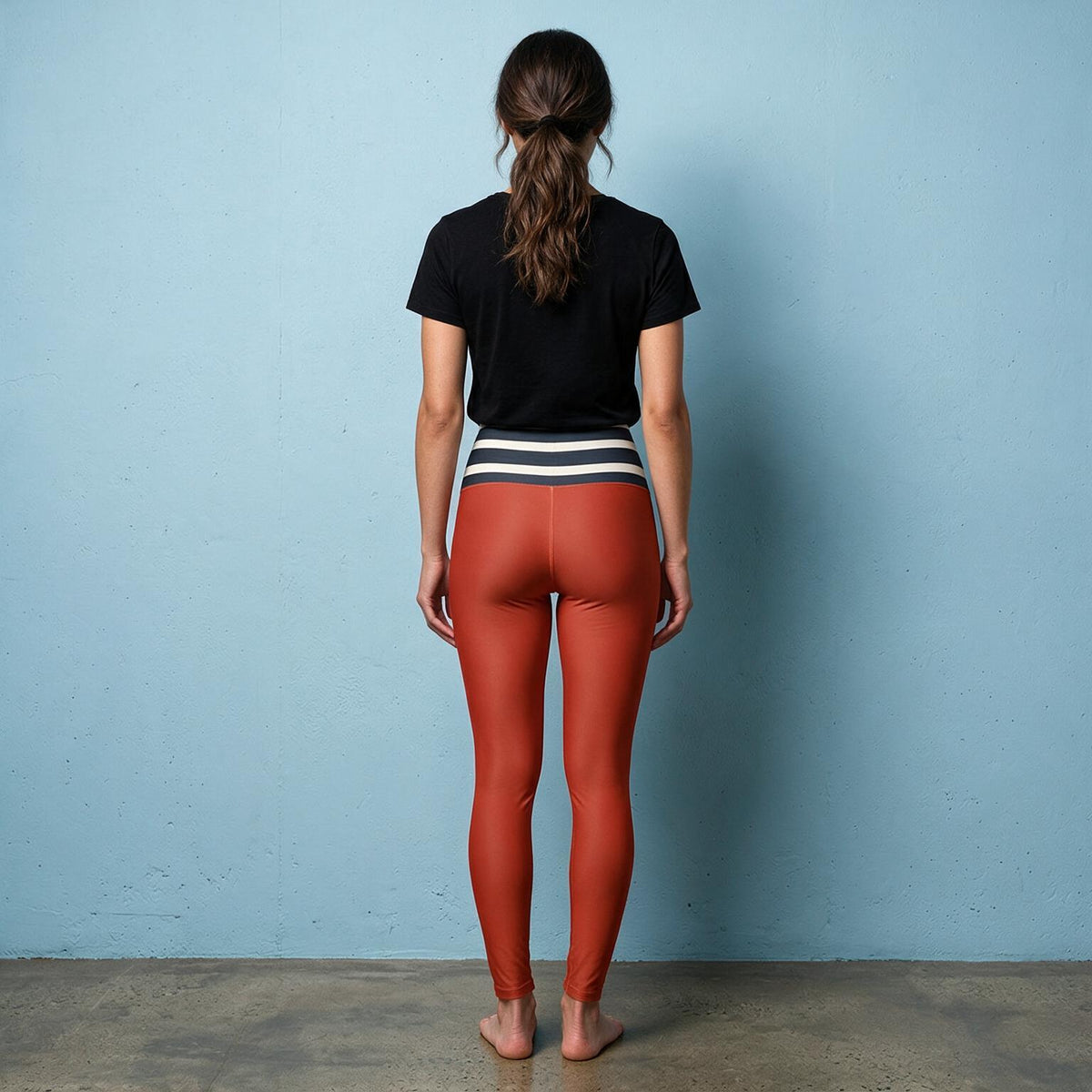 A woman stands with her back to the camera wearing high-waisted rust-orange leggings with a black and white striped waistband, a black t-shirt, and barefoot on a concrete floor against a light blue wall.