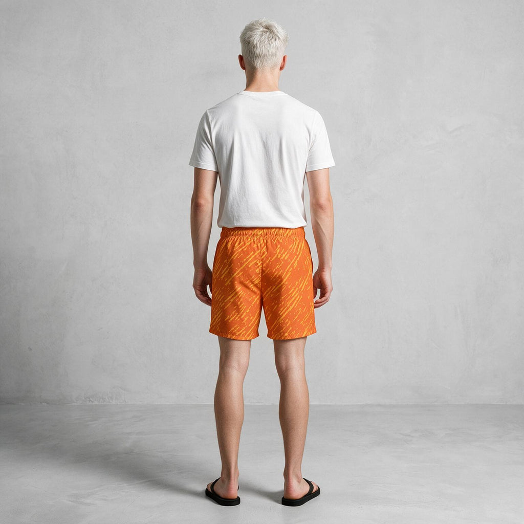 A man viewed from behind wearing a white t-shirt, bright orange patterned swim trunks, and black flip-flops, standing against a light gray textured studio wall.