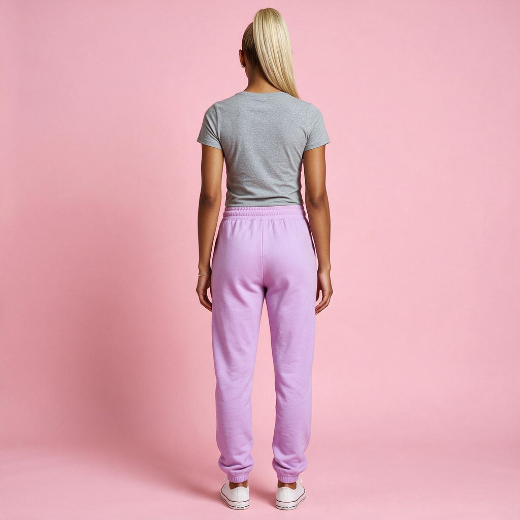 A woman viewed from behind wearing light purple sweatpants, a gray t-shirt, and white sneakers against a solid pink studio background.