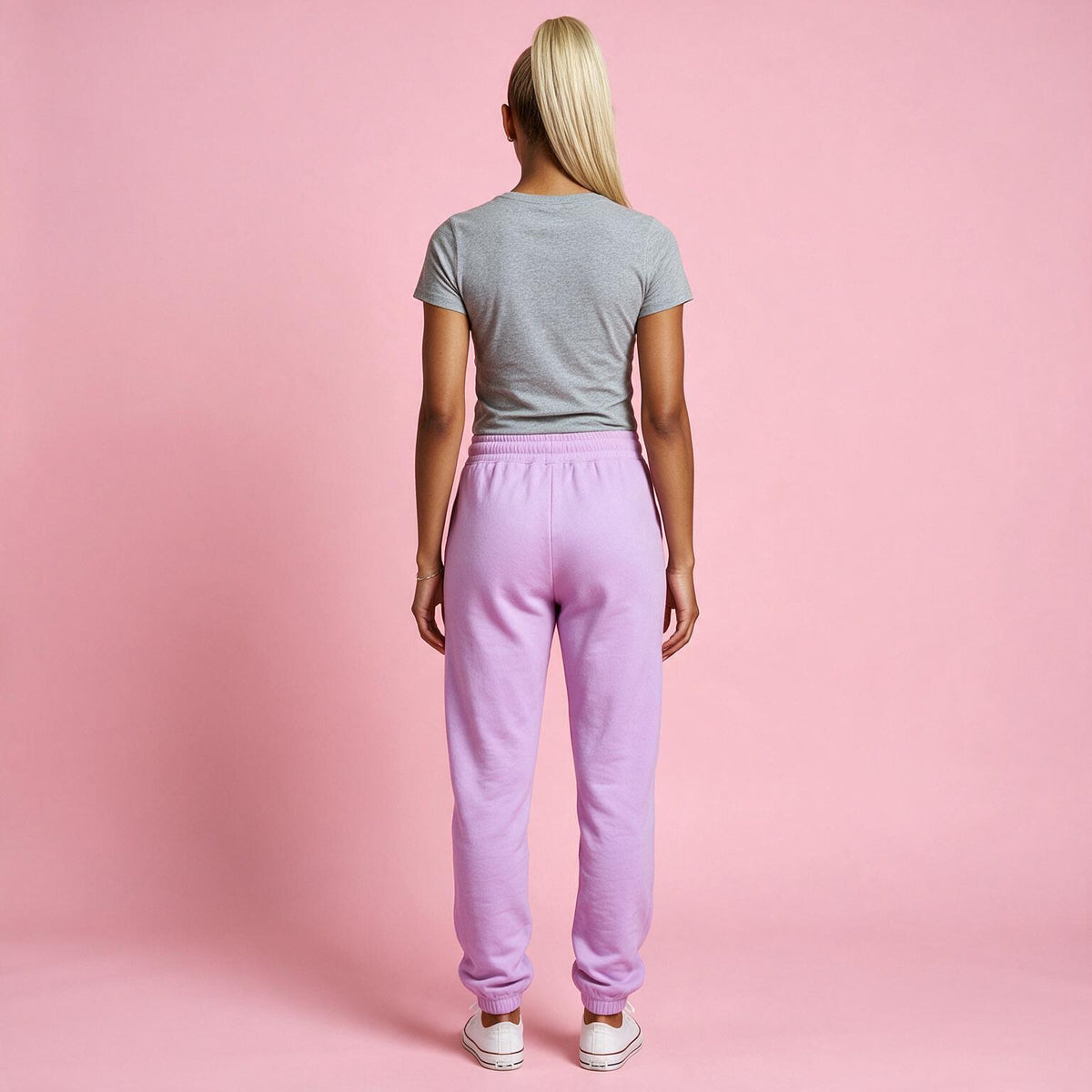 A woman viewed from behind wearing light purple sweatpants, a gray t-shirt, and white sneakers against a solid pink studio background.