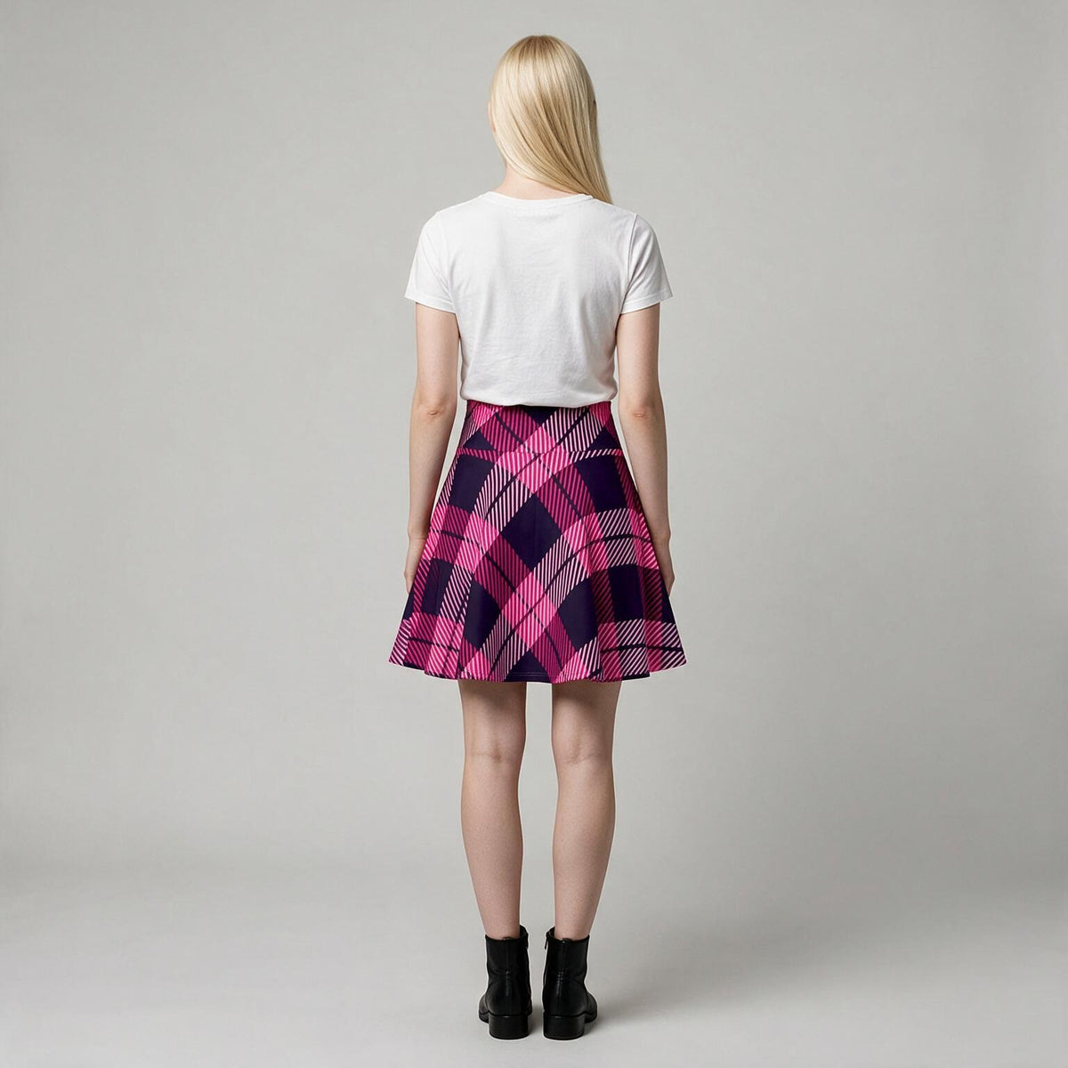 A woman viewed from behind wearing a white short-sleeve crop top and a pink and purple plaid pleated mini skirt, paired with black ankle boots, against a light gray studio background.