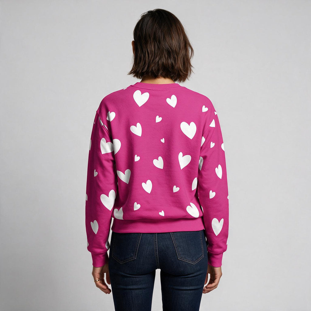 A woman viewed from behind wearing a bright pink crewneck sweatshirt with a white heart pattern, paired with dark blue jeans, against a plain light gray background.