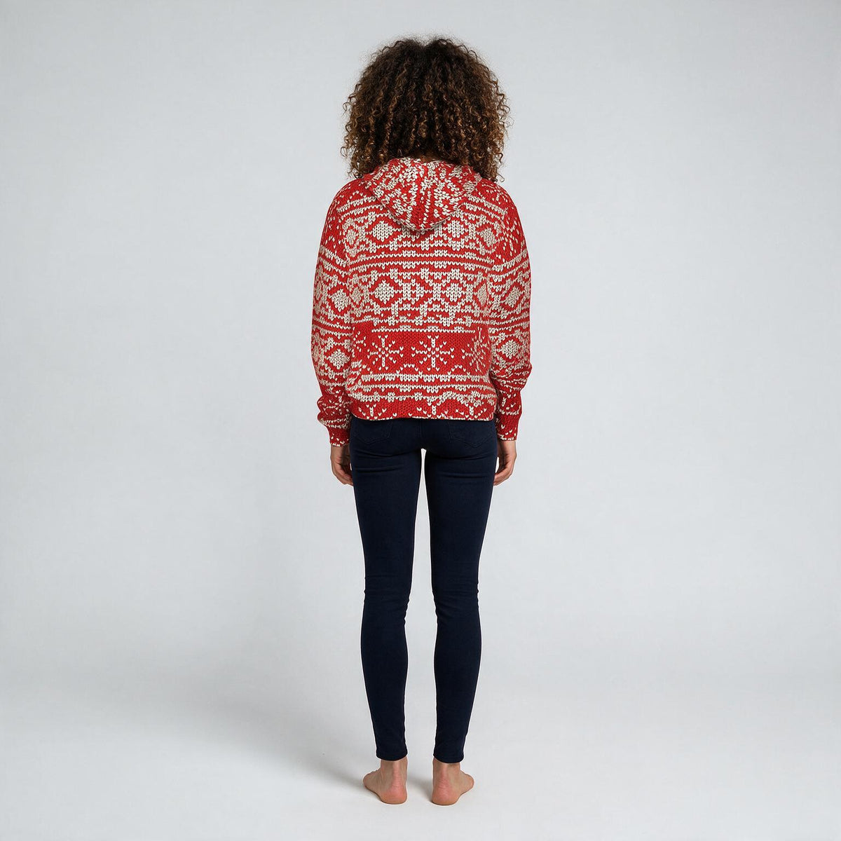A woman viewed from behind wearing a red and white patterned knit hoodie and dark navy leggings, standing against a light gray studio background.