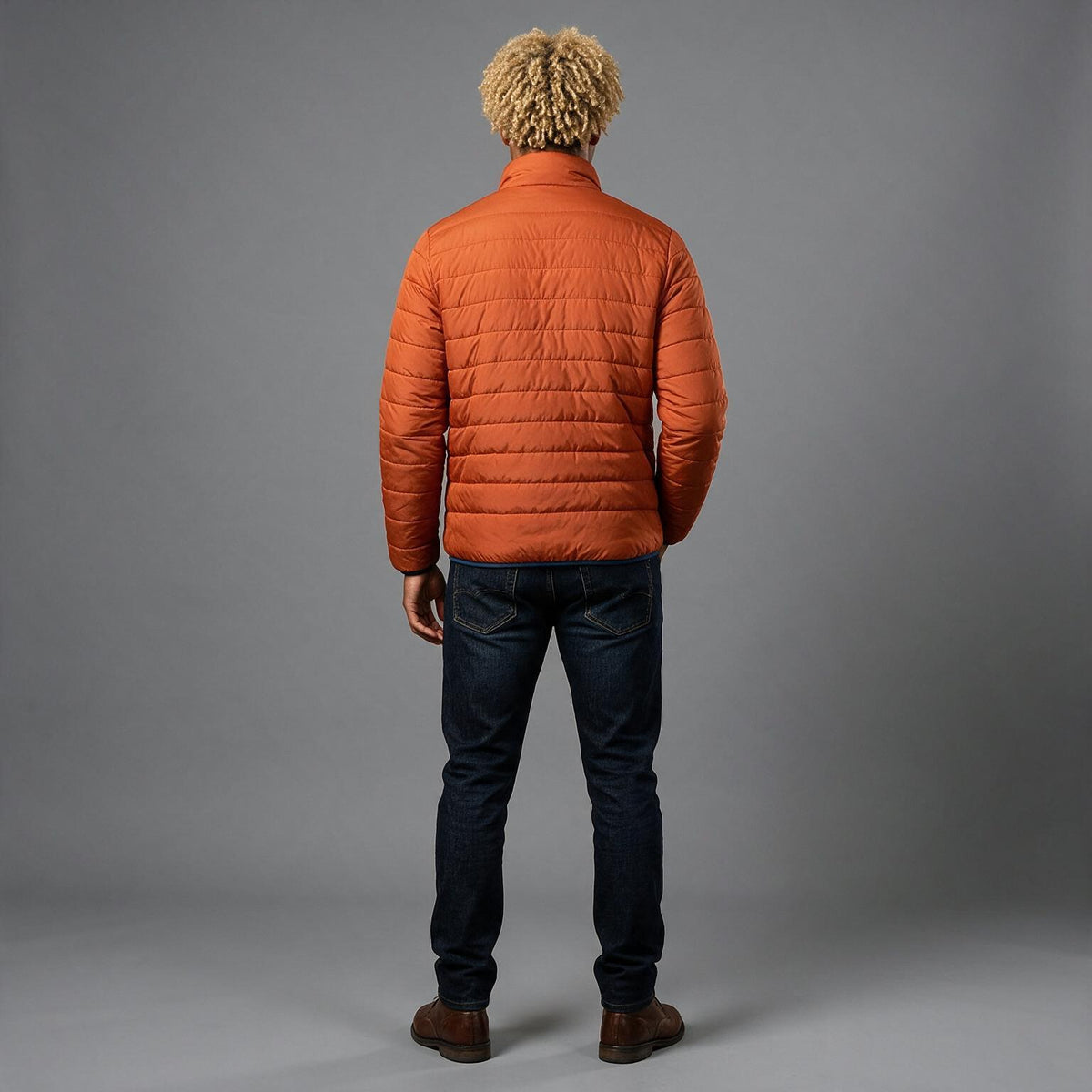 A man viewed from behind wearing an orange quilted puffer jacket, dark blue jeans, and brown shoes, standing against a solid gray studio background.