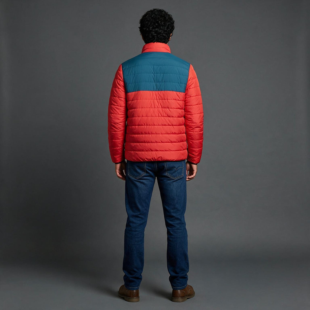A man viewed from behind wearing a color-blocked puffer jacket with a teal blue upper back and shoulders and a bright red lower back, sleeves, and hem, paired with blue jeans and brown shoes, against a dark gray studio background.