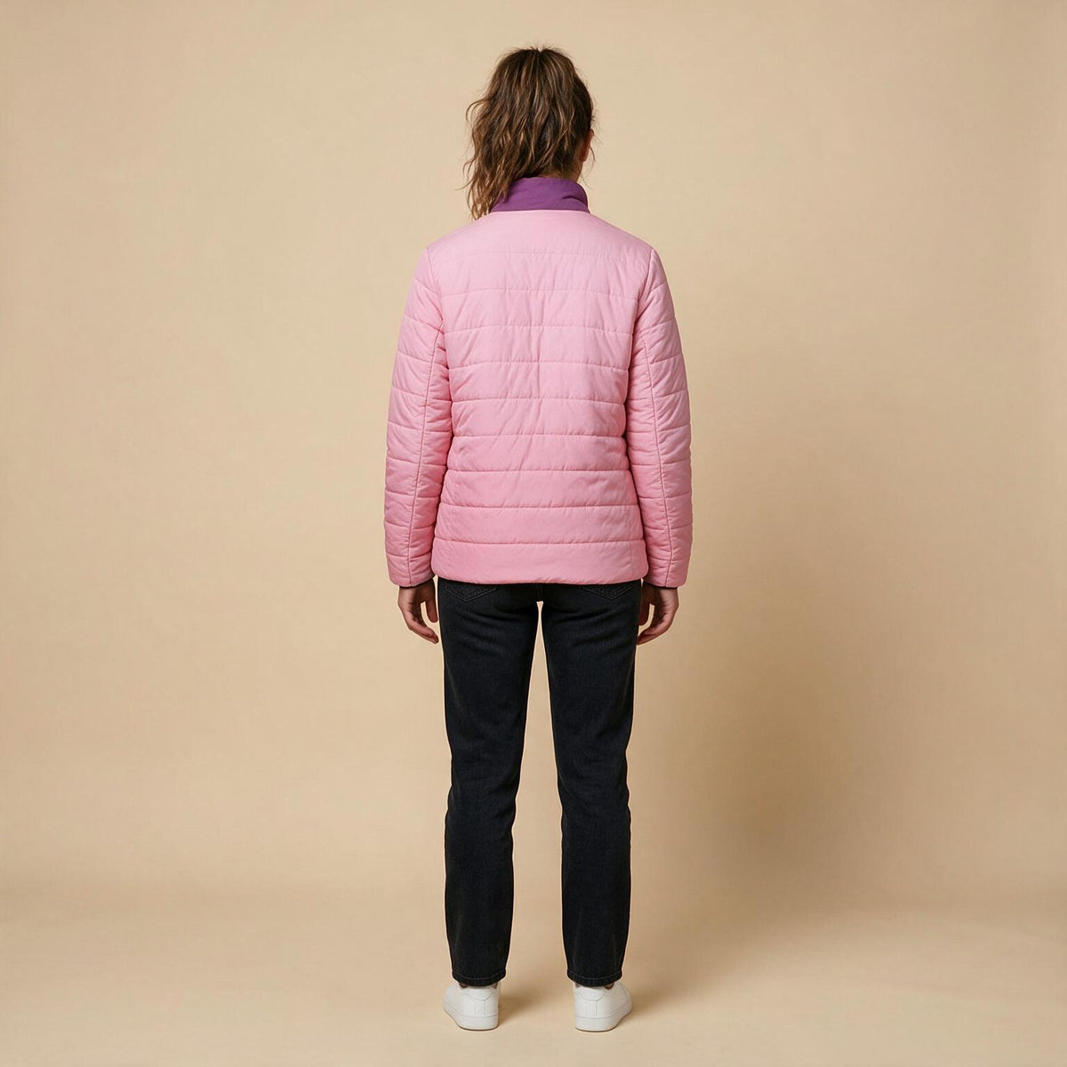 A woman viewed from the back wearing a light pink quilted puffer jacket with a contrasting purple collar, paired with dark pants and white shoes, against a solid beige studio background.